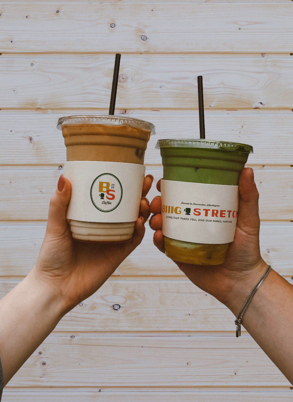 Two hands holding iced coffee drinks in clear plastic cups with black straws, with a light wooden wall background.
