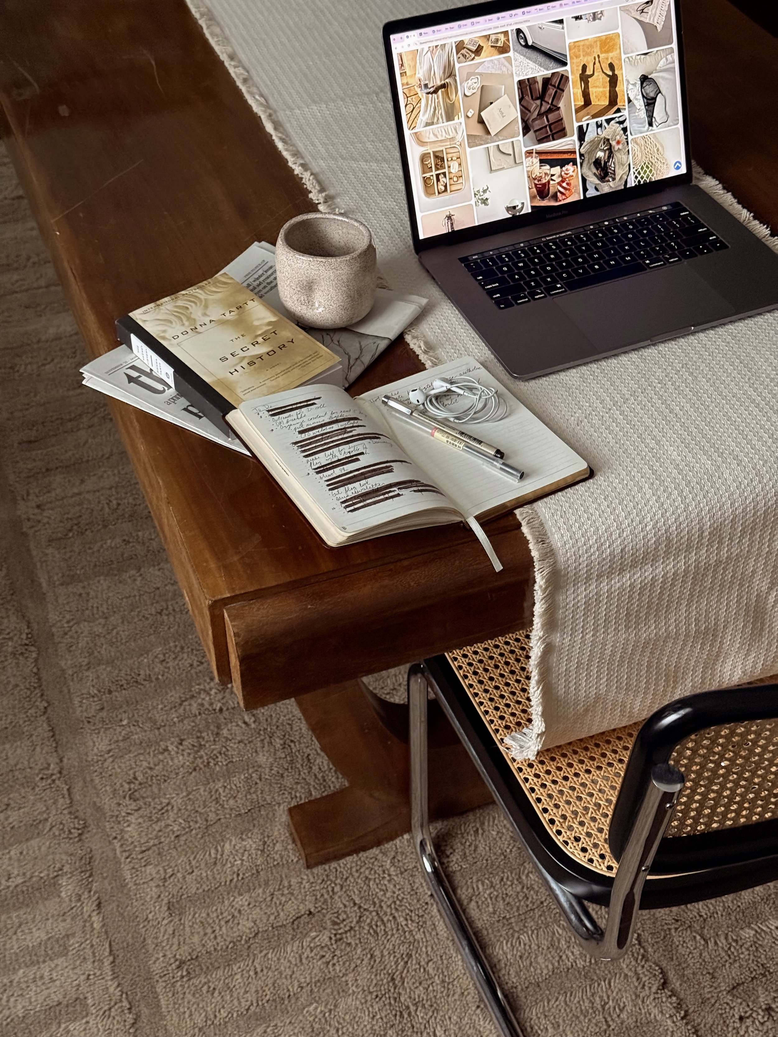 A workspace with a wooden table holding a MacBook laptop, a notebook with handwritten notes, a pen, AirPods, a book titled 'The Secret History' by Donna Tartt, and a ceramic mug. A beige cloth covers part of the table, and a woven chair is underneath the table. The area is carpeted with a neutral-colored rug.