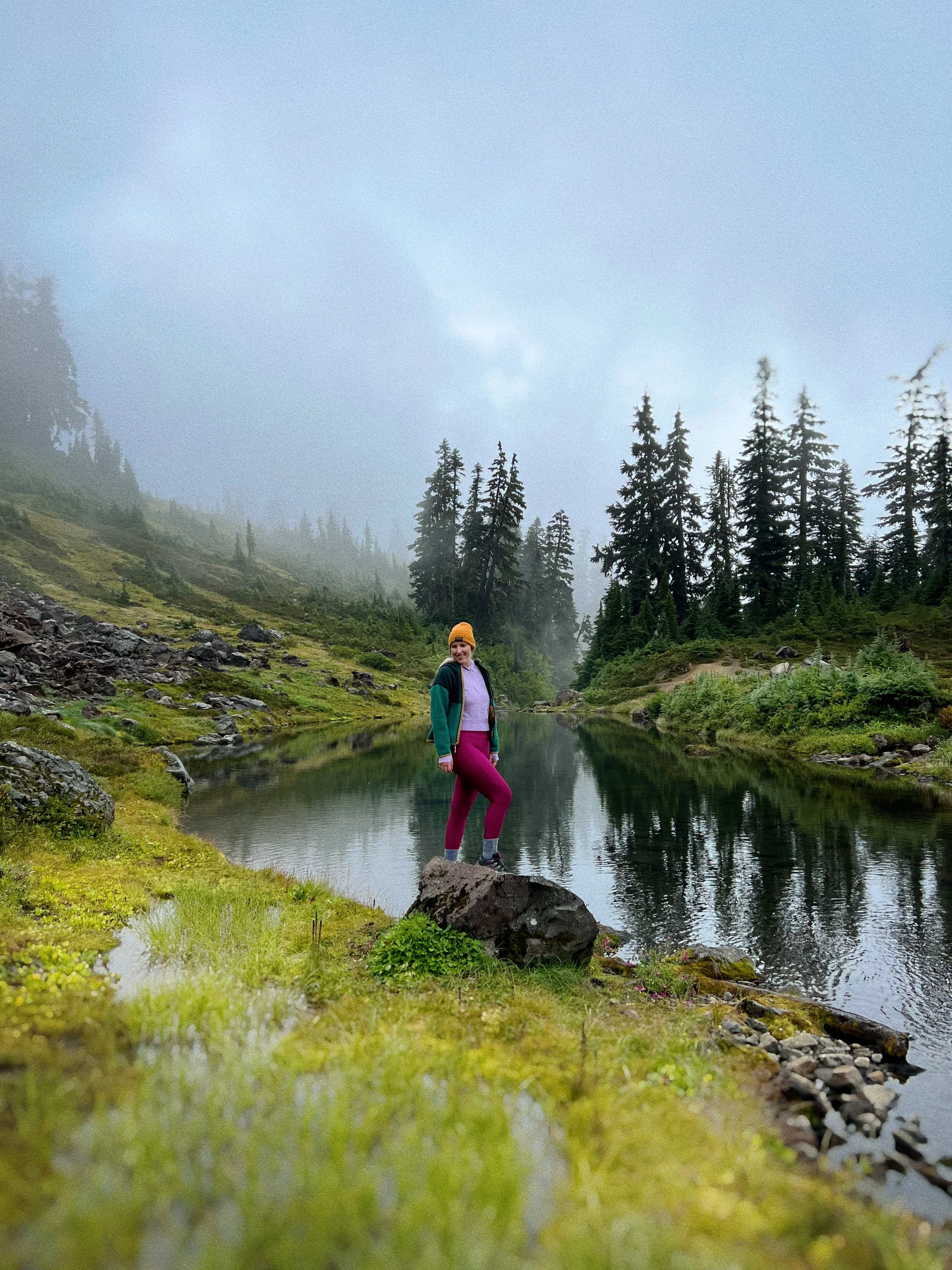 A woman standing on a rock by a peaceful mountain lake surrounded by dense evergreen trees and foggy weather.