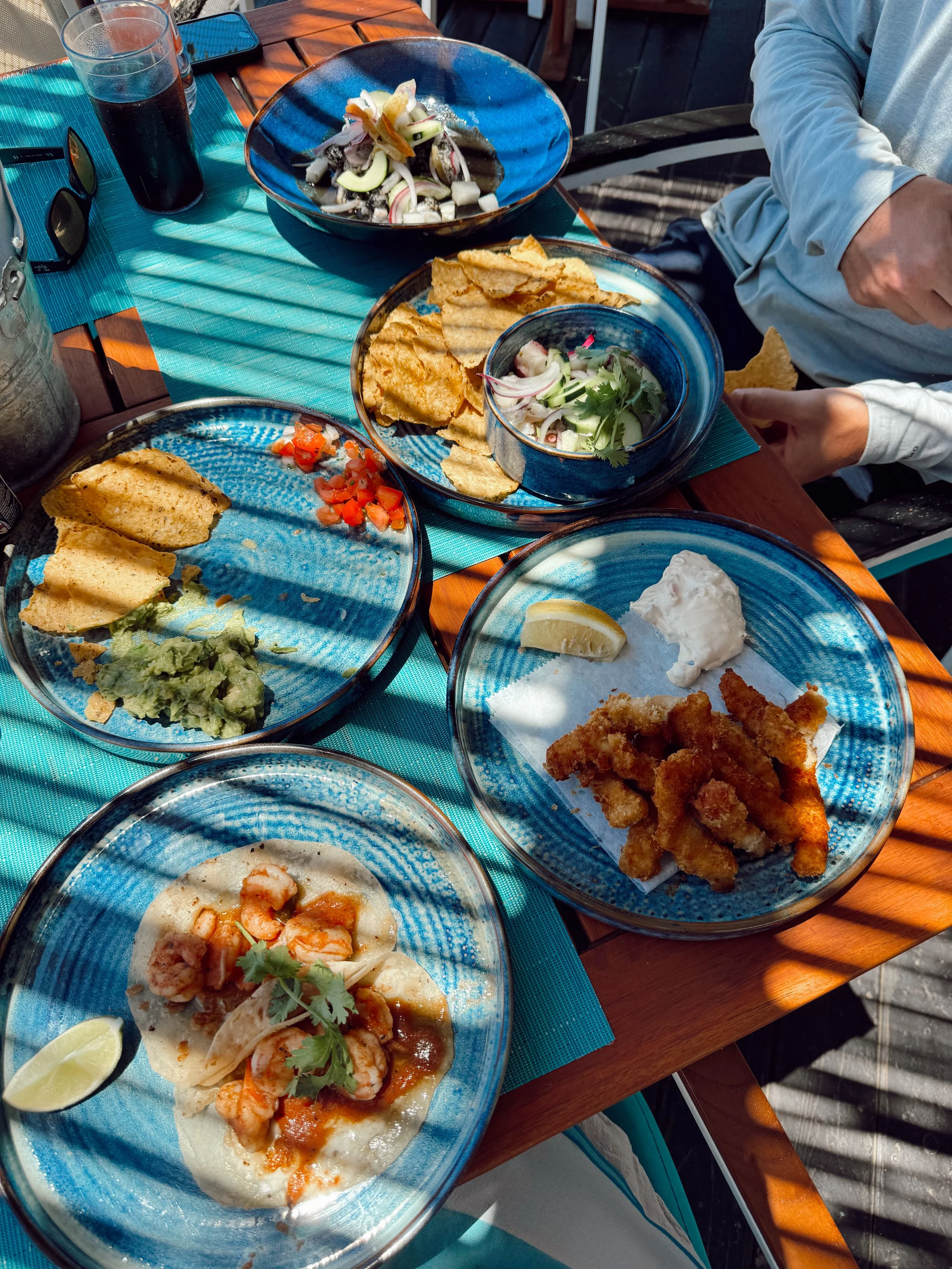 A table with various plates of Mexican food, including tacos, chips, guacamole, salsa, and seafood dishes, on a blue tablecloth