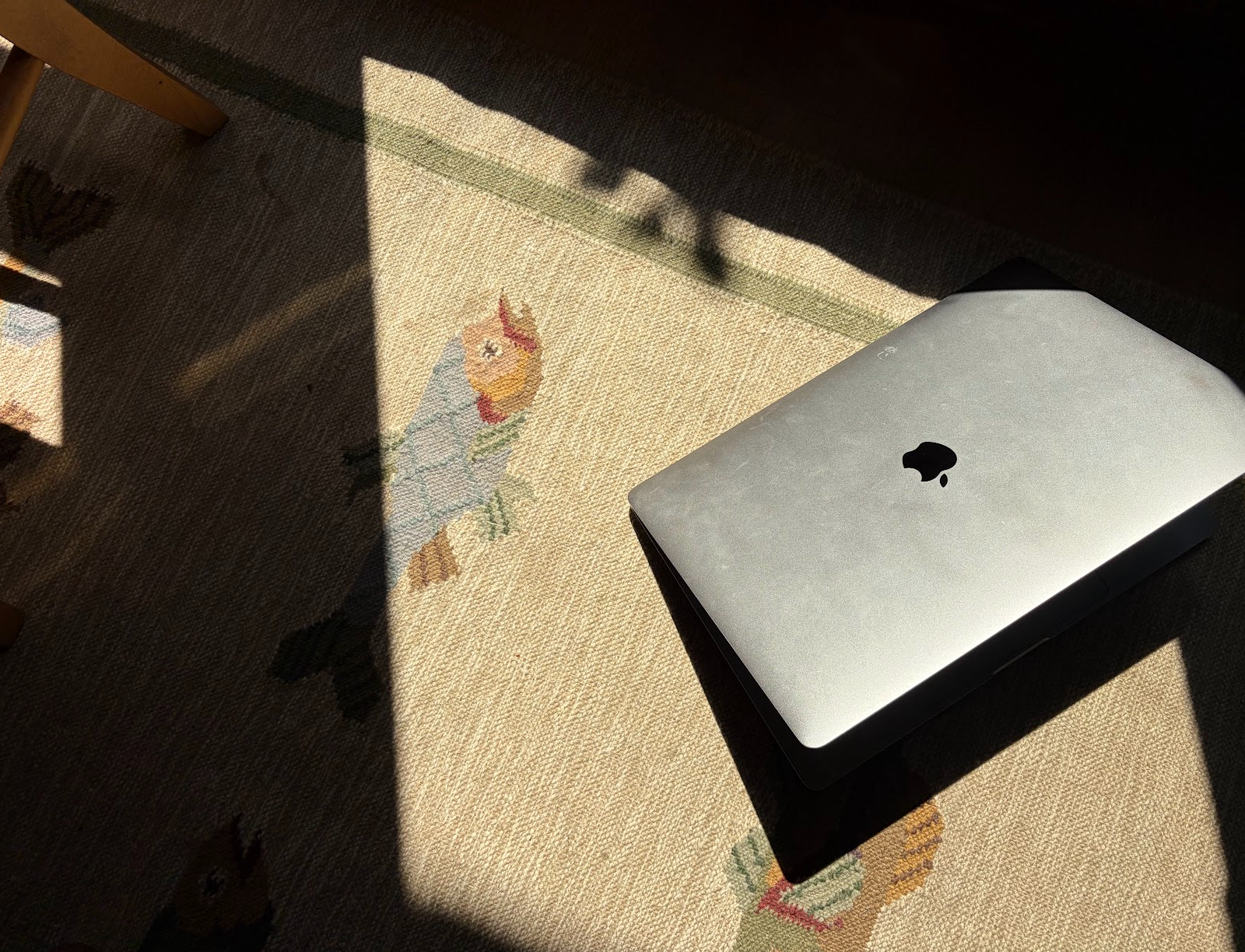A closed silver MacBook laptop on a colorful embroidered tablecloth with floral patterns, partially illuminated by sunlight, with shadows cast over the surface.