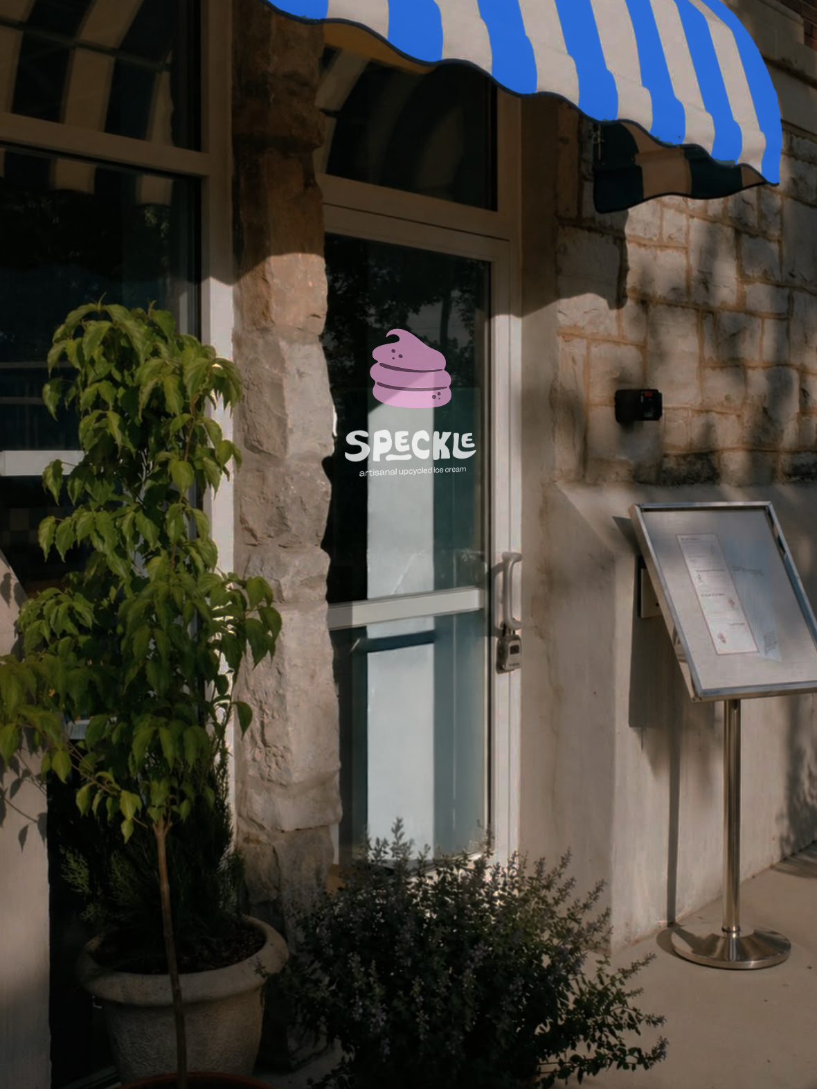 Glass door with a Speckle ice cream shop sticker, surrounded by potted plants, with a menu stand on the sidewalk outside and a stone wall and window in the background.