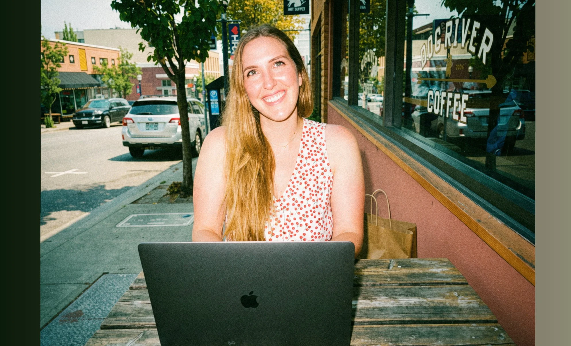 A young woman with long blonde hair smiling while sitting outside at a table with a laptop in front of her. She is near a coffee shop with a large window that has the words 'DO', 'RIVER', and 'COFFEE' painted on it. The background shows a street with parked cars and trees.