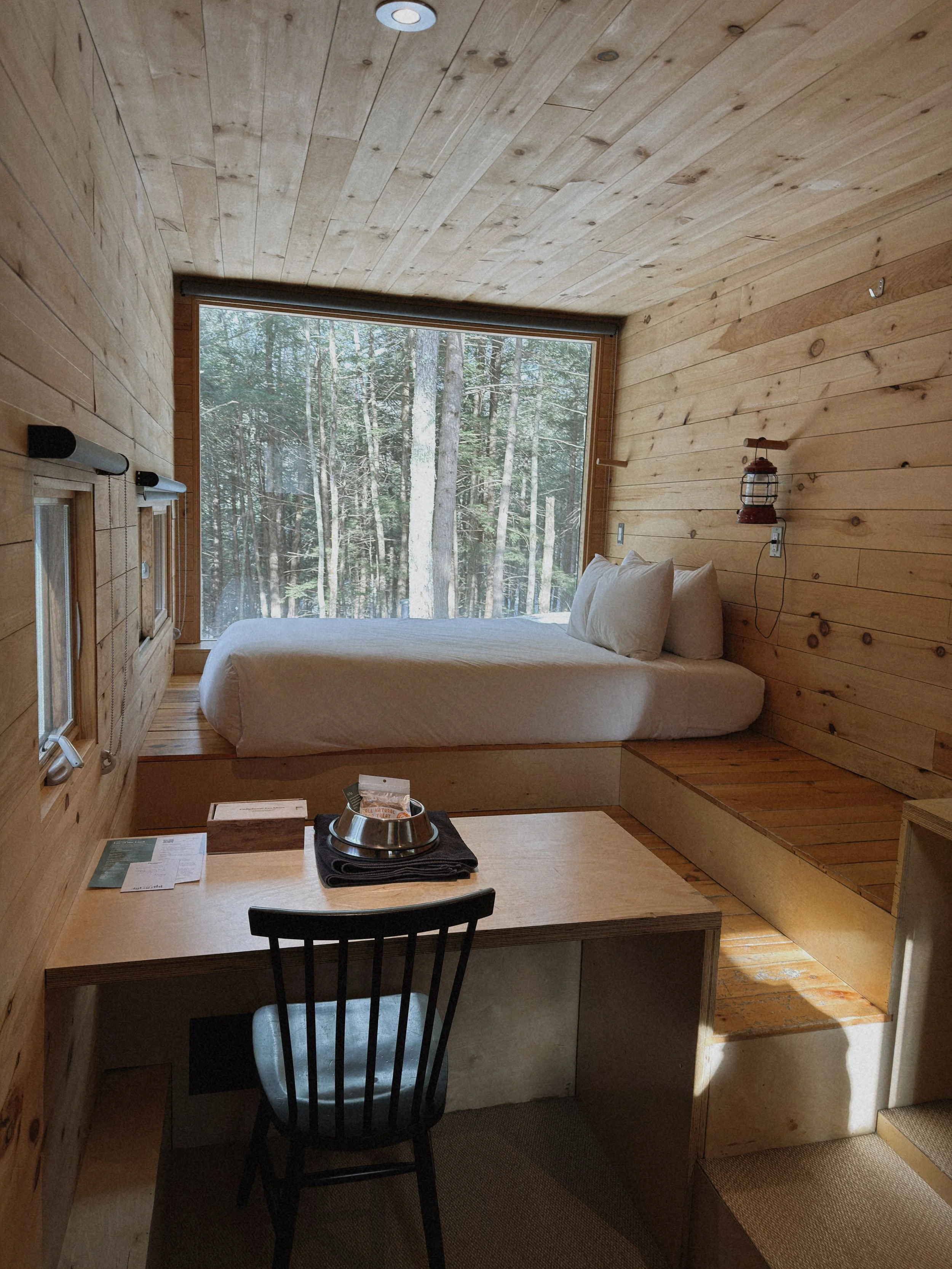 Small cozy room with wooden walls and ceiling, large window overlooking a forest, bed with white bedding, black chair at a desk, and a small wood-burning stove mounted on the wall.