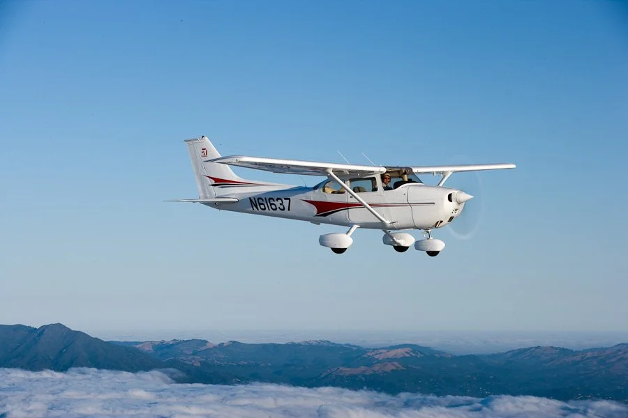 Small white aircraft flying above mountain landscape with a clear blue sky.