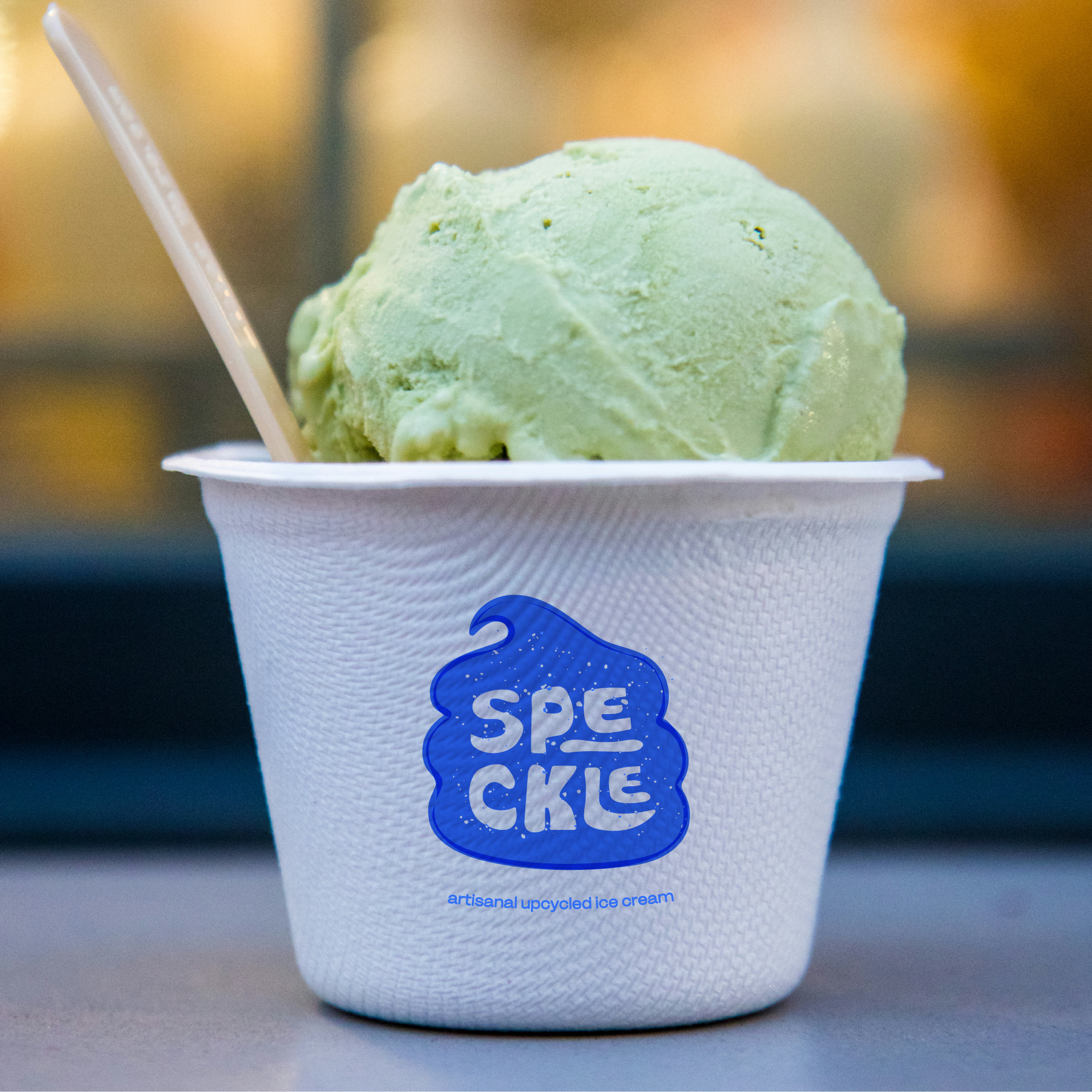A cup of green artisanal ice cream in a white paper cup with a blue logo that reads "SPE CICLE."