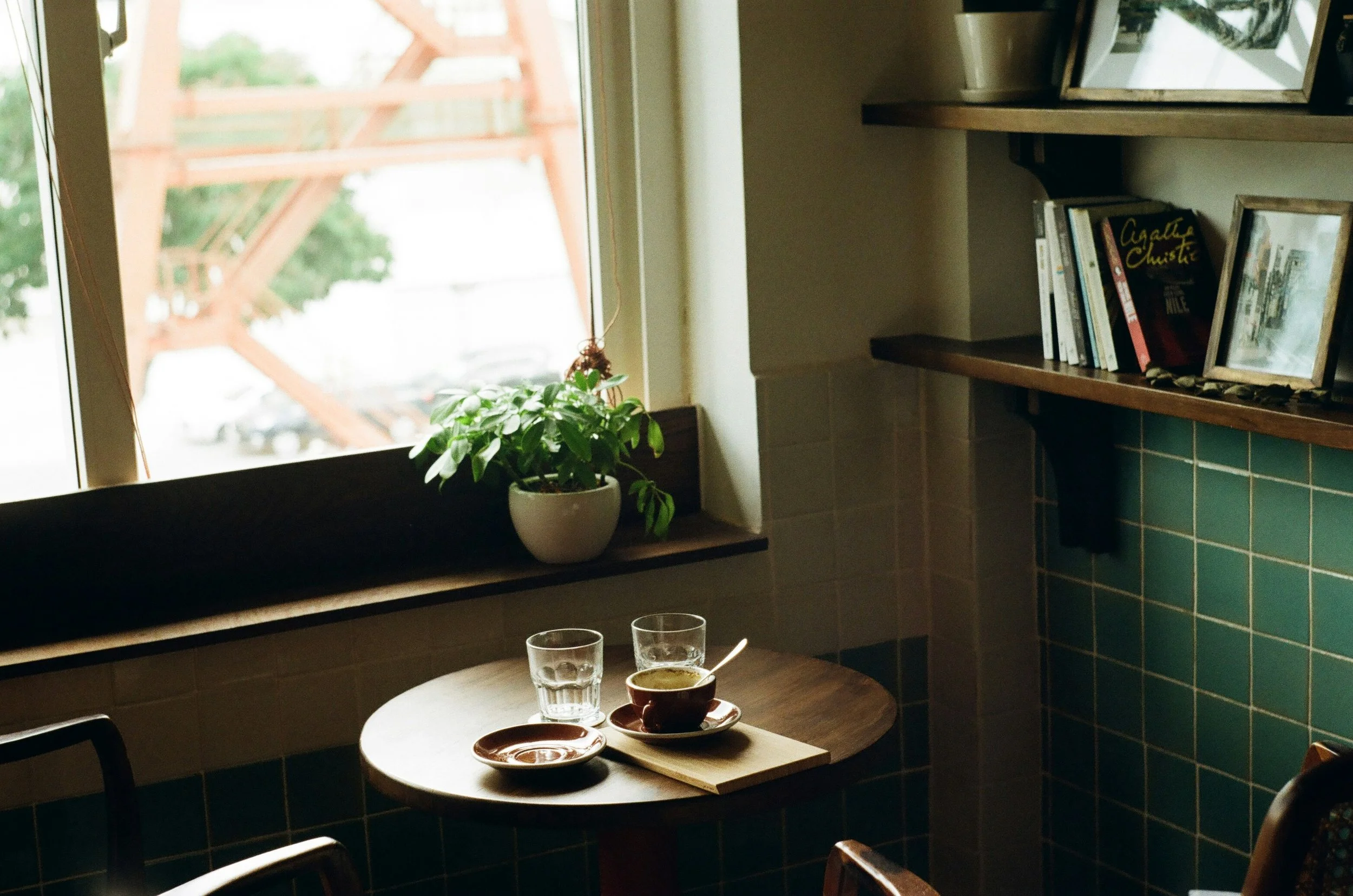 Cozy cafe interior with a window, potted plant, bookshelf with books, and table set with coffee cups, glasses of water, and cutlery.