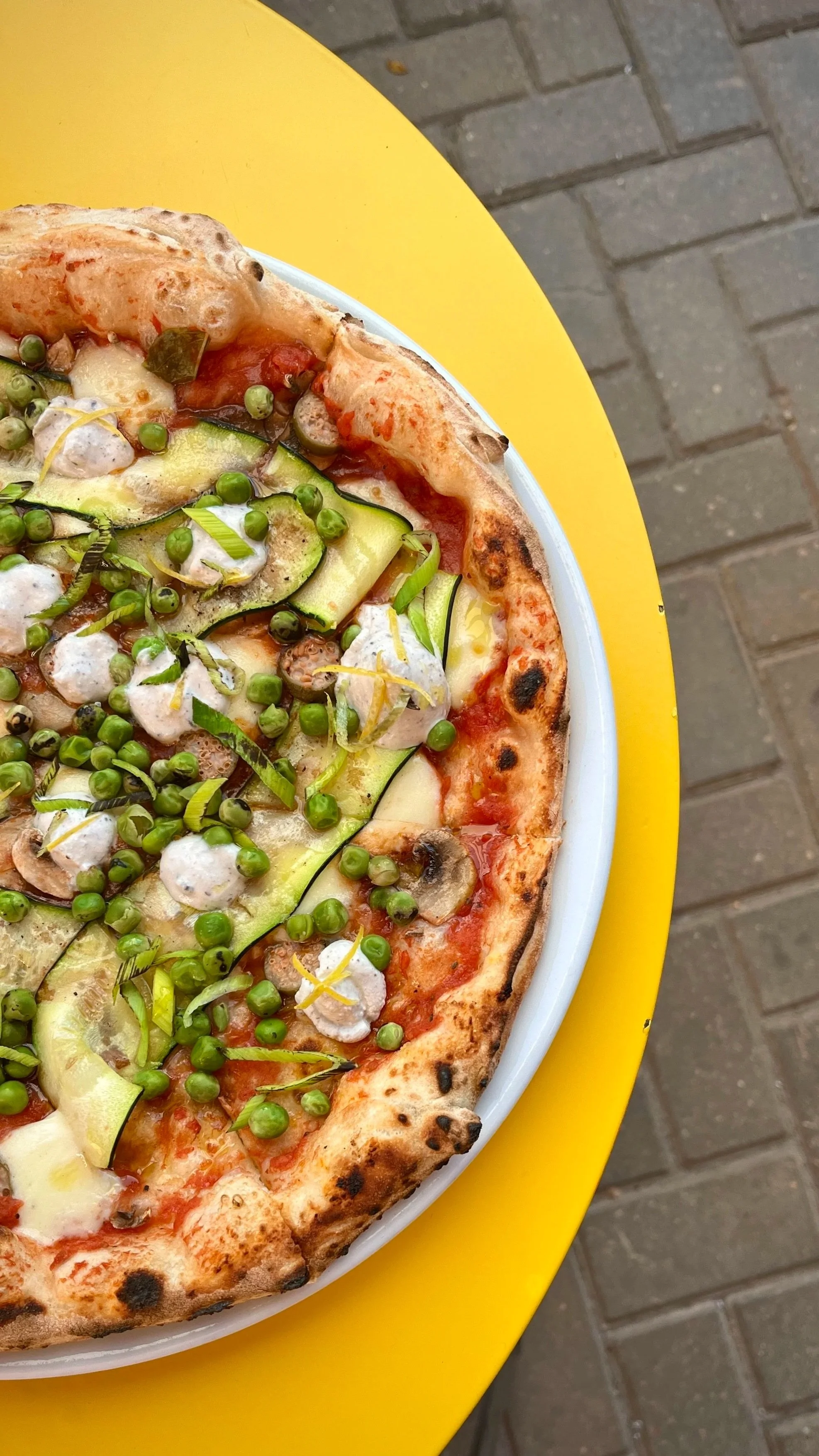 Close-up of a pizza with vegetables and cheese on a white plate, placed on a yellow table. The background shows a brick pavement.