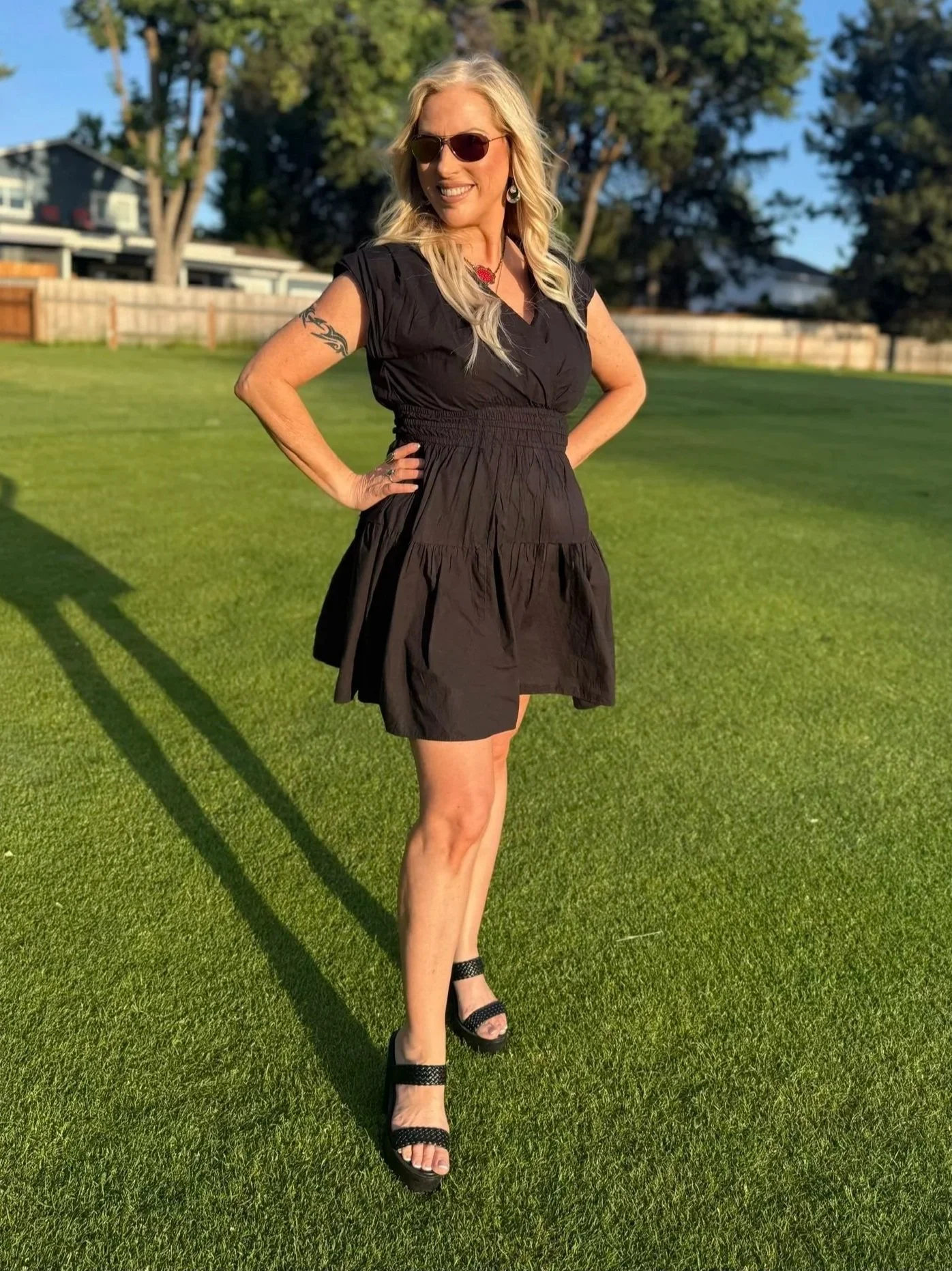 A woman with blonde hair, wearing sunglasses, a black dress, and black high heels, standing on a grassy area with trees and houses in the background during sunset.