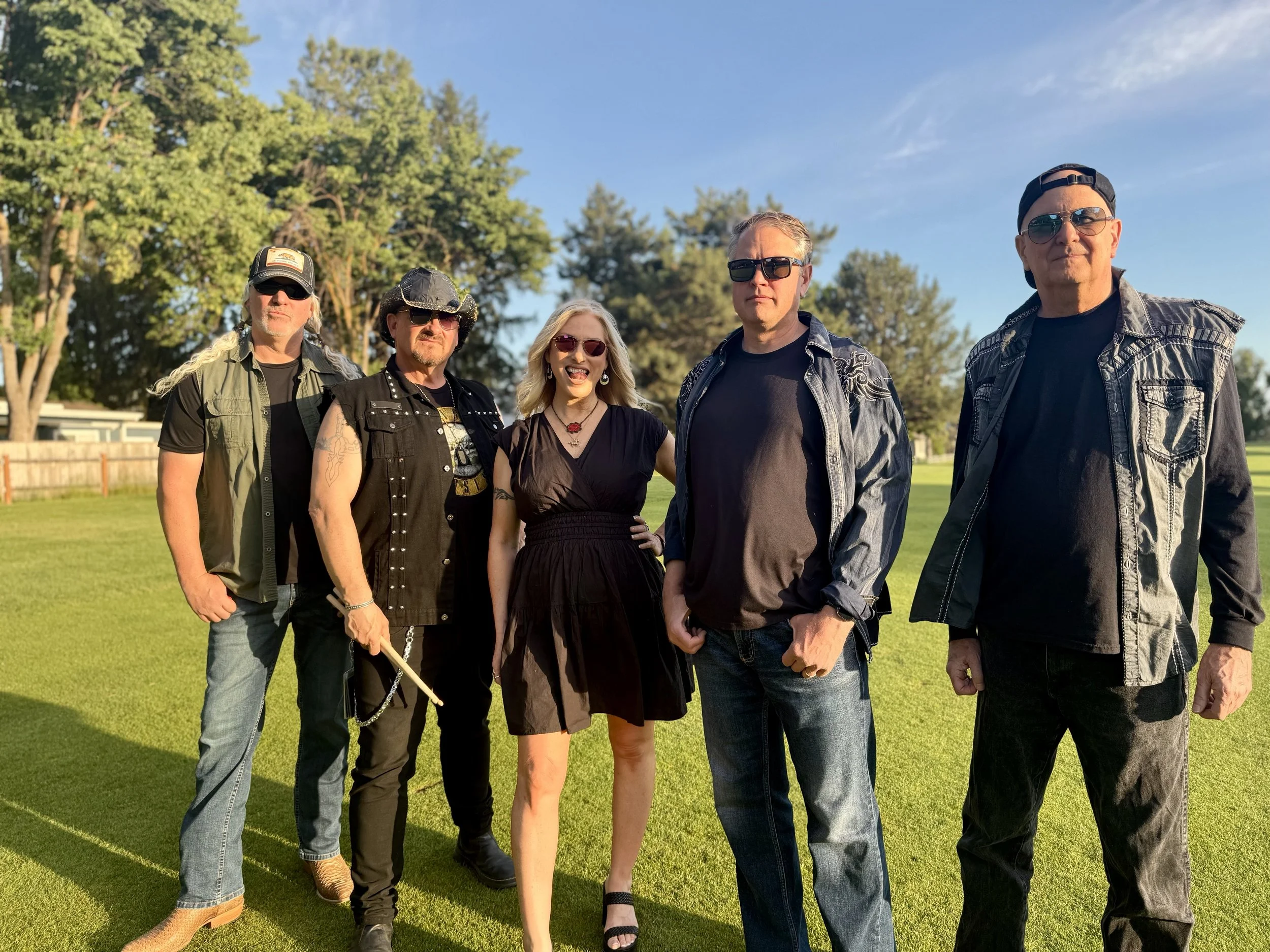 Stylus Down, a band of five people, standing outdoors on a sunny day with trees and a blue sky in the background.
