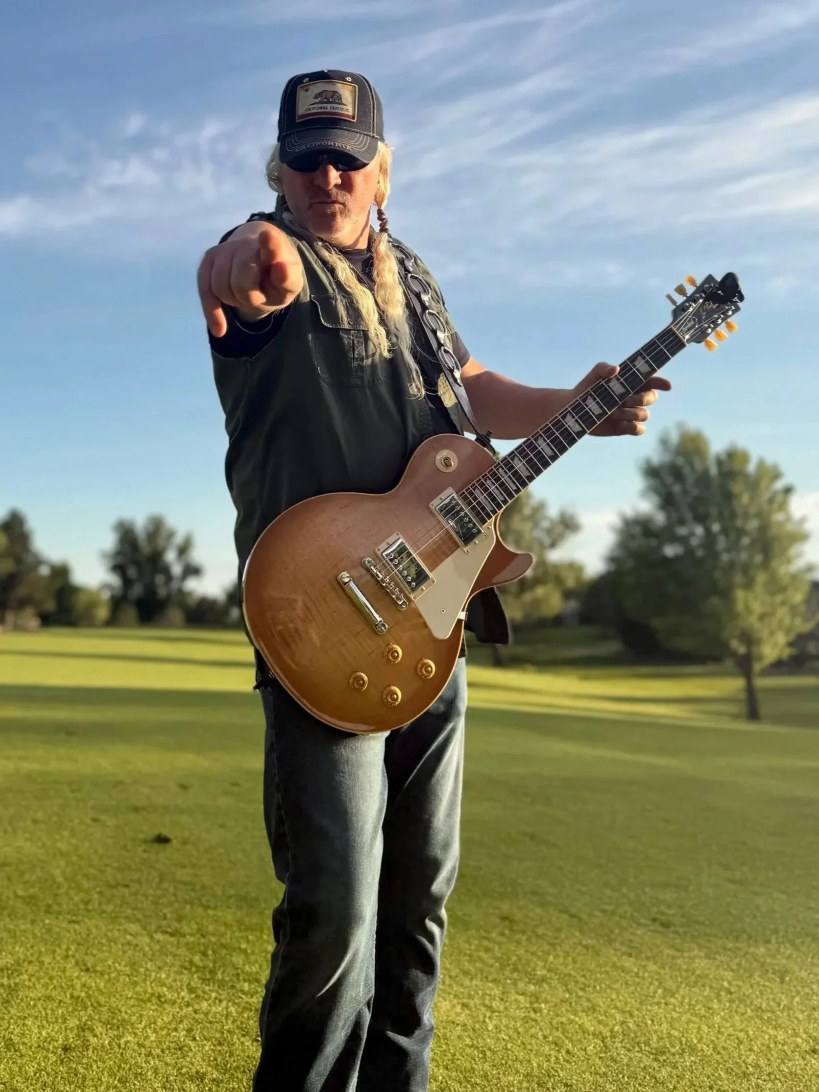 A man with long blonde hair, wearing a black cap, black sunglasses, and black vest, points at the camera while holding an electric guitar. He stands outdoors on a grassy field with trees and a blue sky in the background.