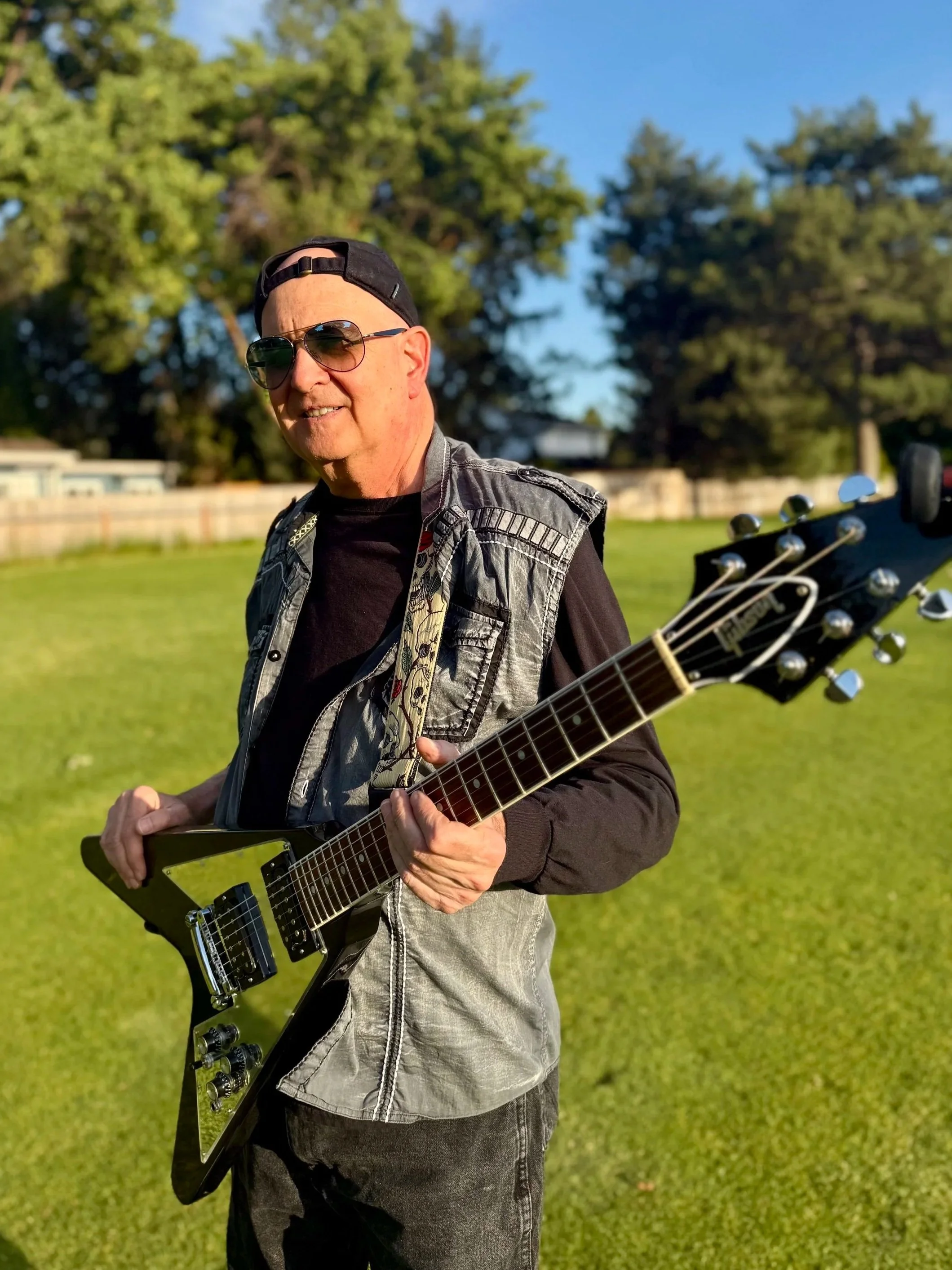 A man wearing sunglasses and a backwards cap playing an electric guitar outdoors on a grassy field with trees in the background.