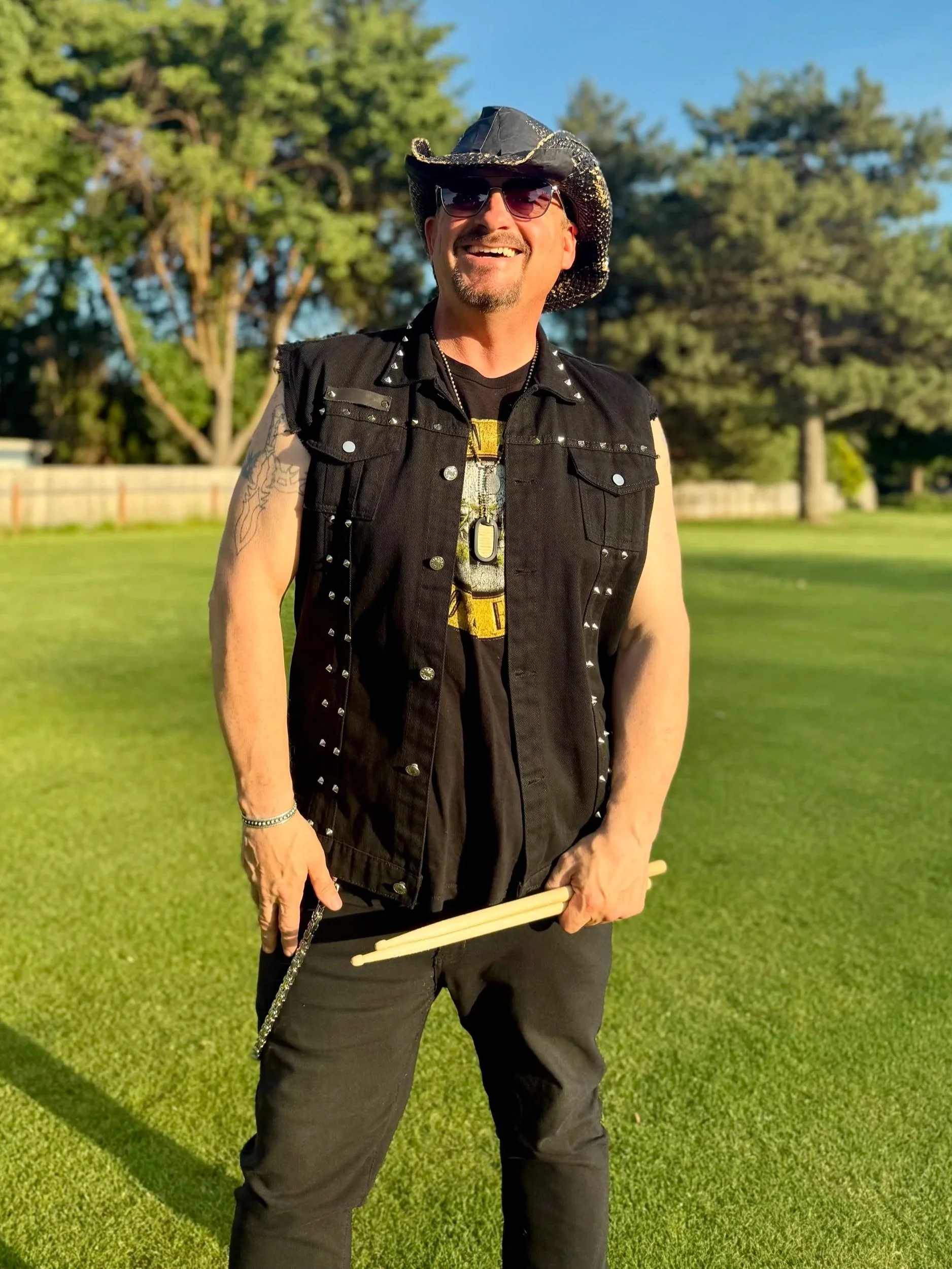 A man smiling outdoors in a grassy park, wearing a black sleeveless vest with silver studs, a cowboy hat, sunglasses, and holding drumsticks.