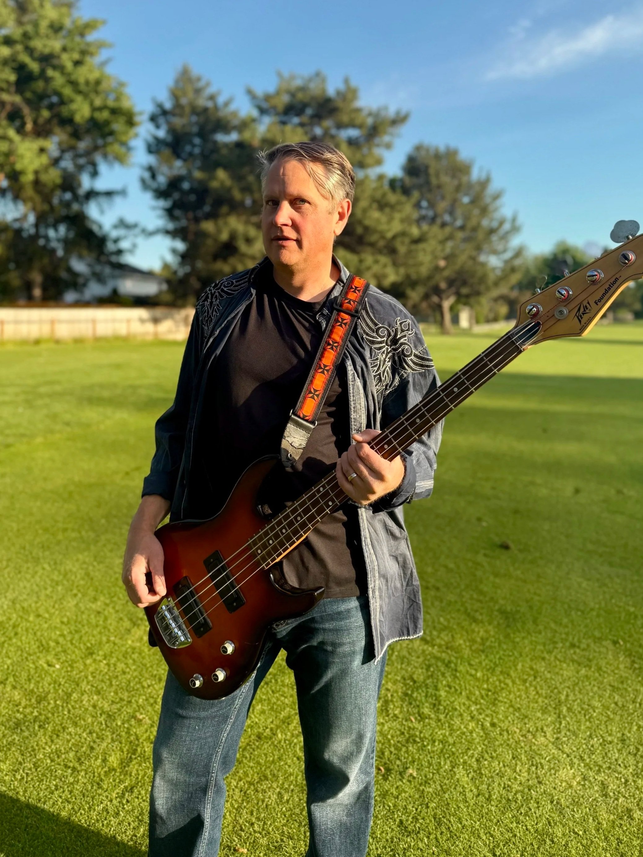 A man standing on a grassy field holding a bass guitar, wearing a dark shirt and jeans, with trees and a clear blue sky in the background.