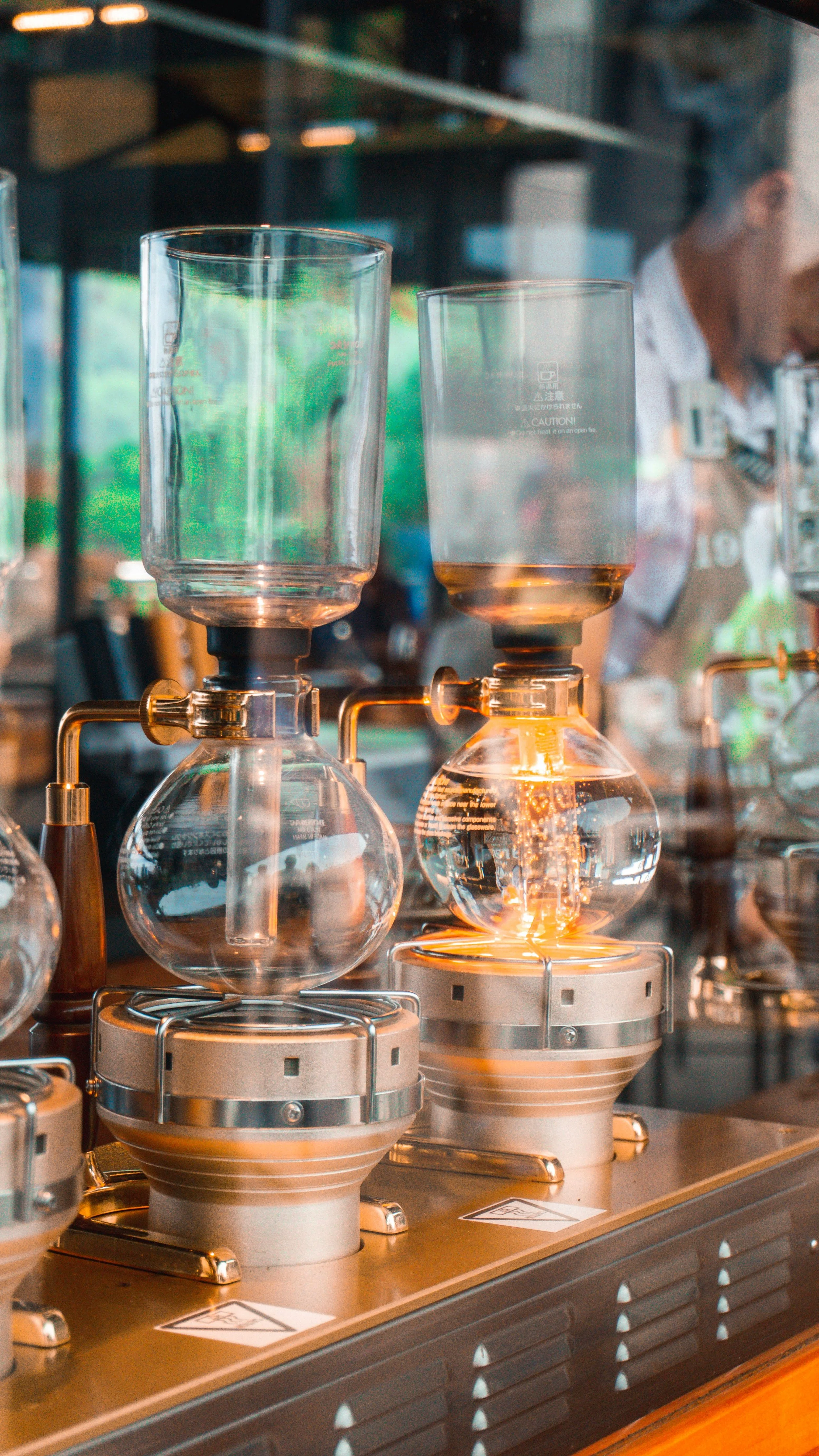 Glassware filled with liquid connected to a coffee brewing machine, with a person in the background blurred.