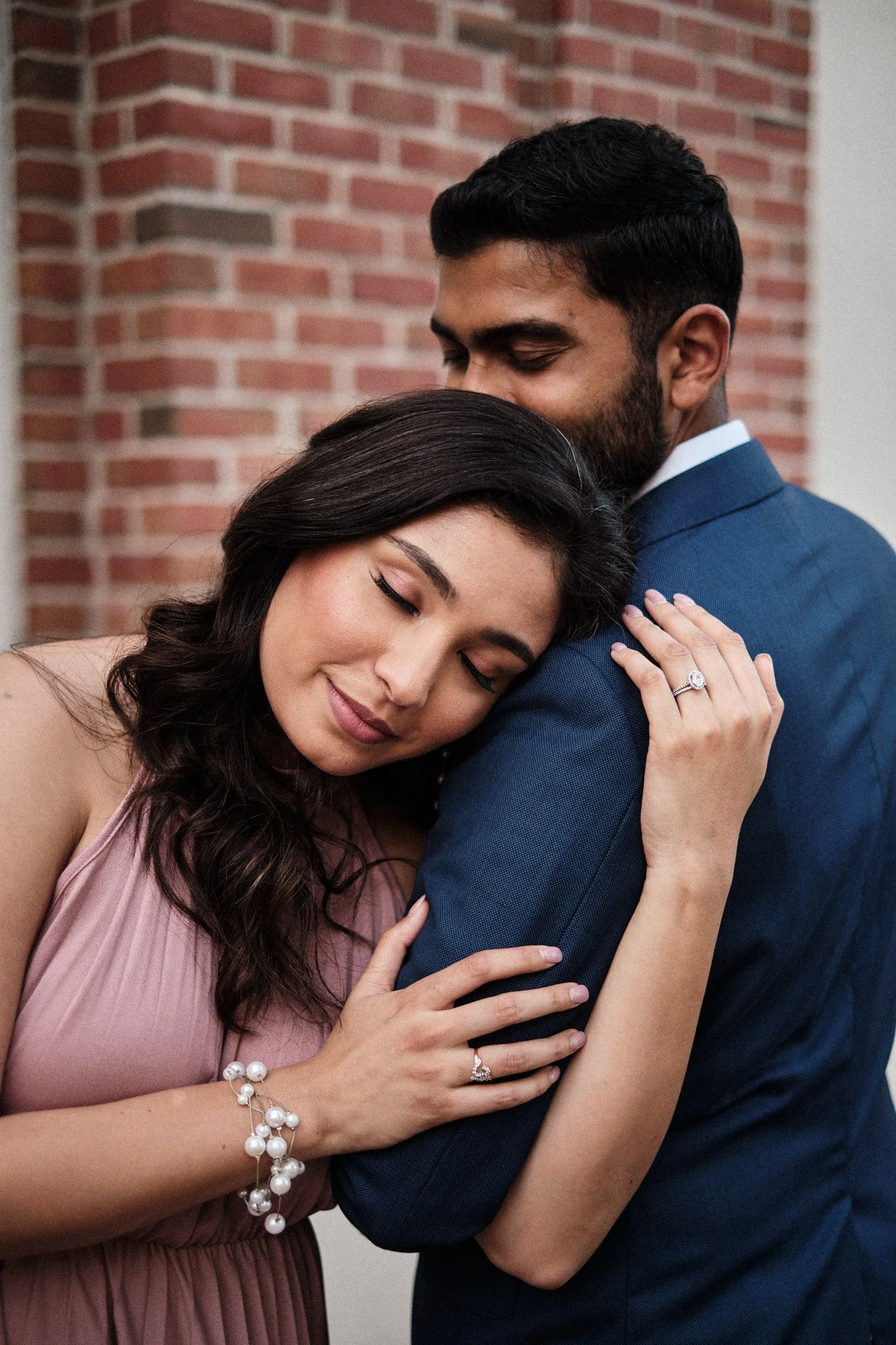 A woman and a man sharing a hug, with the woman resting her head on the man's shoulder and smiling. The woman is wearing a pink dress and jewelry, and the man is wearing a blue suit. They are standing in front of a brick wall.