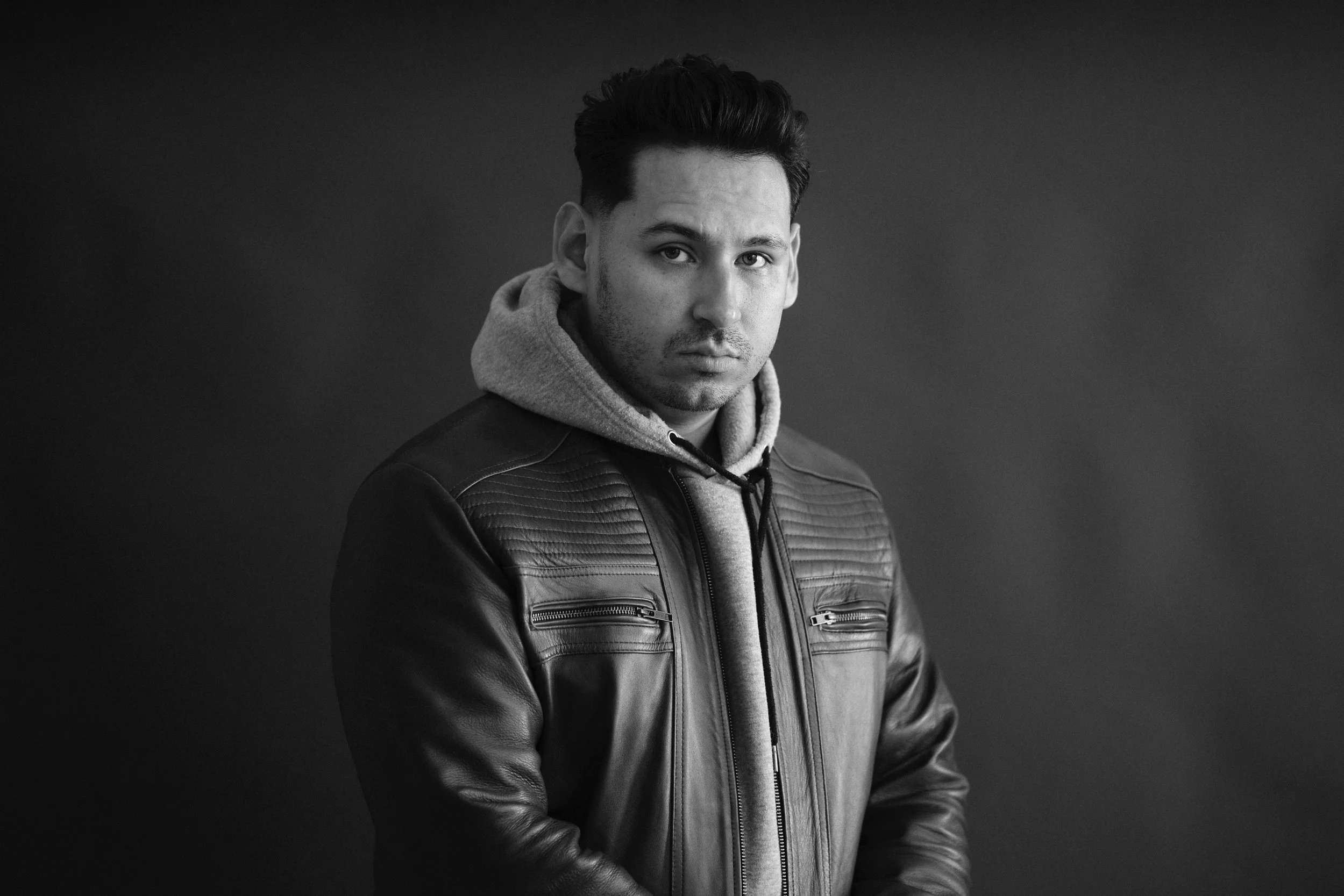 Black and white portrait of a young man with short dark hair, wearing a leather jacket over a hoodie, looking directly at the camera against a dark background.