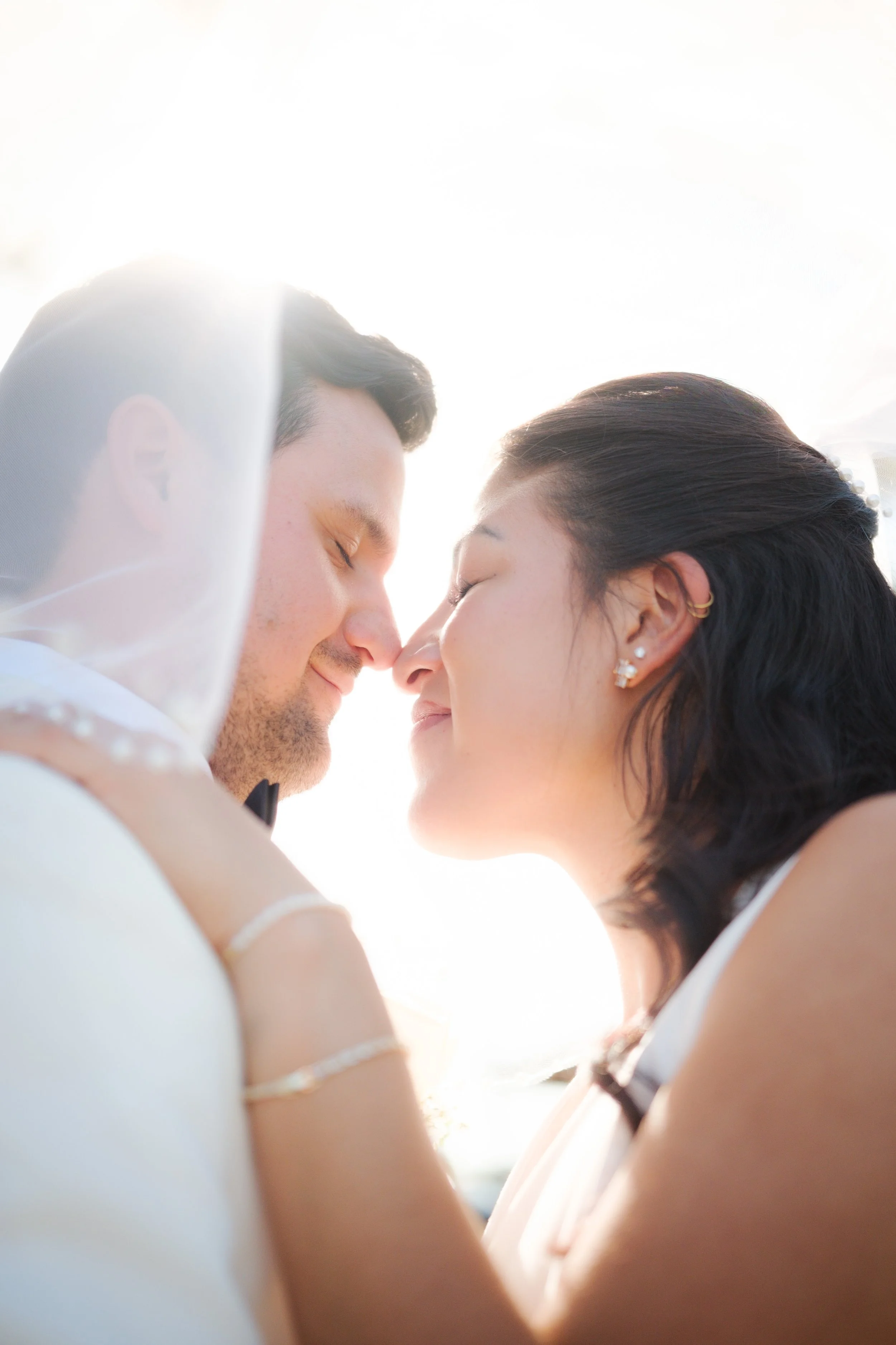 A couple is about to kiss outdoors in bright sunlight, with their foreheads touching and eyes closed.