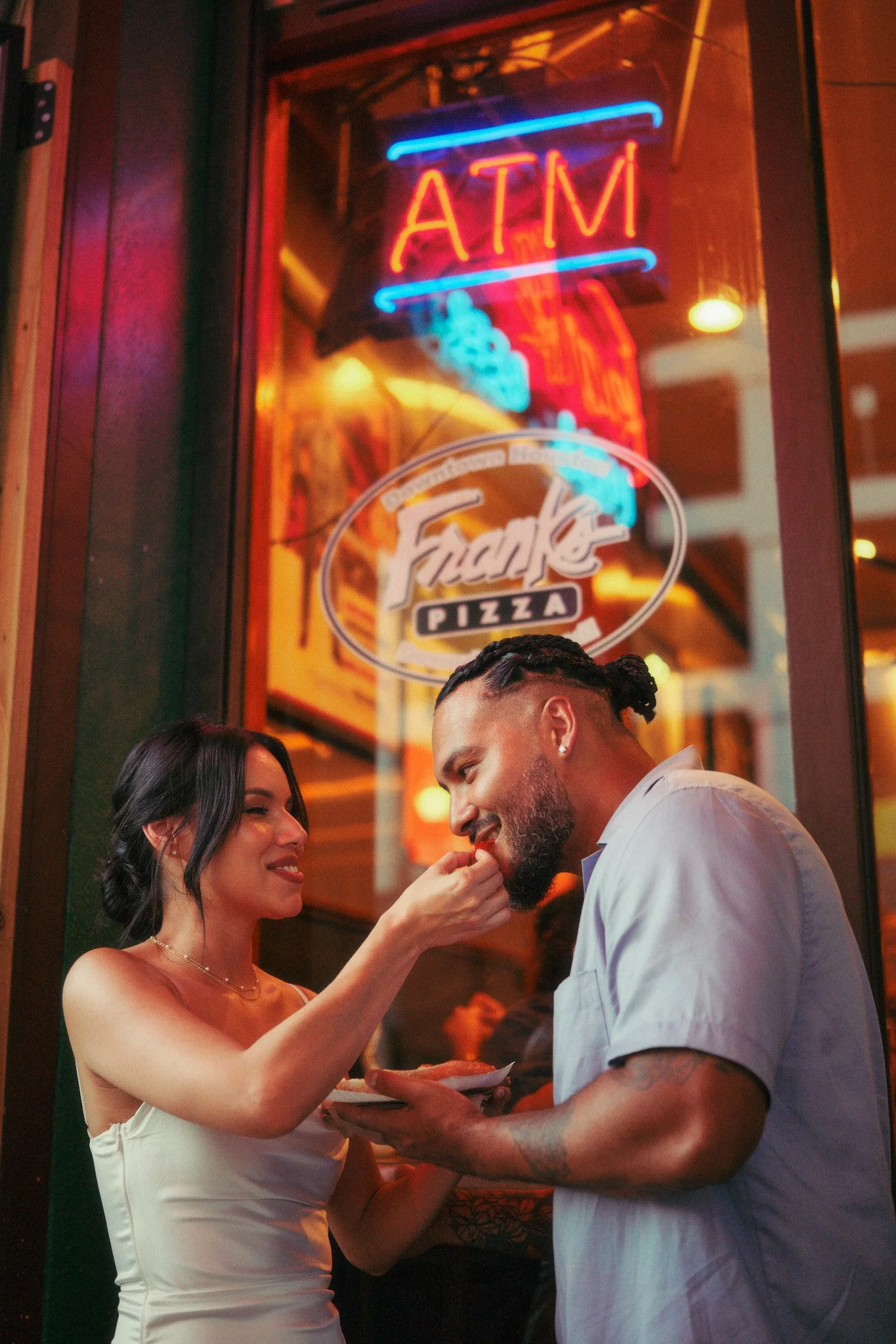 A woman in a white dress is feeding a man in a white shirt with food from a plate, in front of a restaurant or pizza place with neon signs reading ATM and Pizza.