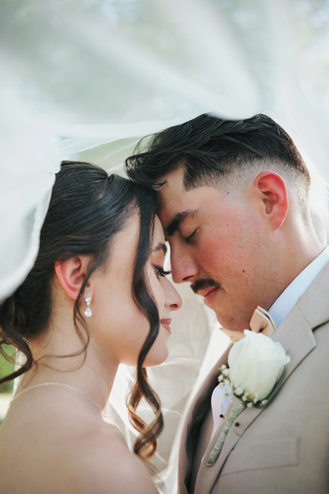 A close-up of a bride and groom with their foreheads touching, eyes closed, during a wedding ceremony, under a canopy or veil.