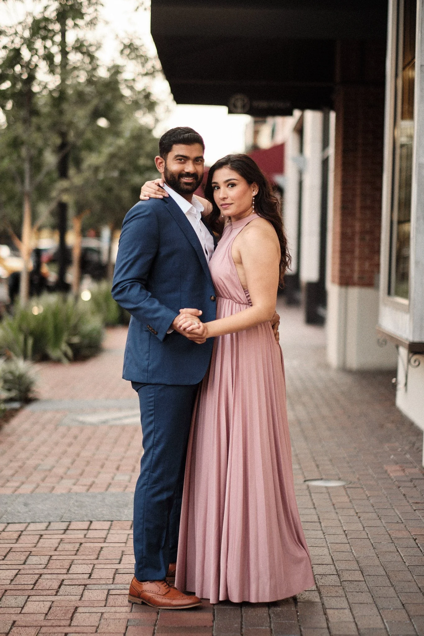 A man and woman dressed in formal attire standing close together outdoors on a brick sidewalk, smiling at the camera. The man is wearing a navy blue suit and brown shoes, and the woman is wearing a long, pink halter-neck gown with earrings. In the ba