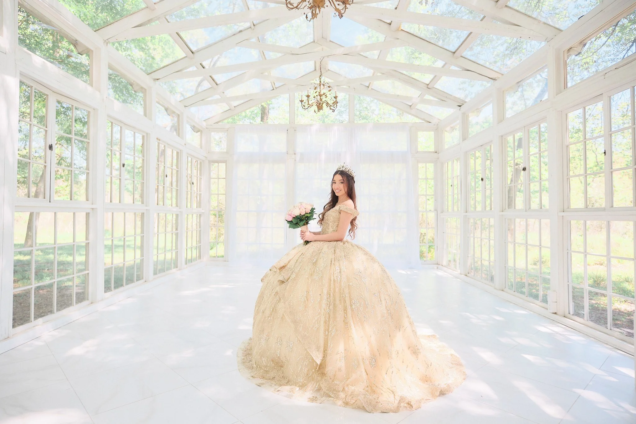 A young woman in a gold ball gown standing inside a bright sunroom with large windows and a glass ceiling, holding a bouquet of pink roses, smiling, wearing a tiara.