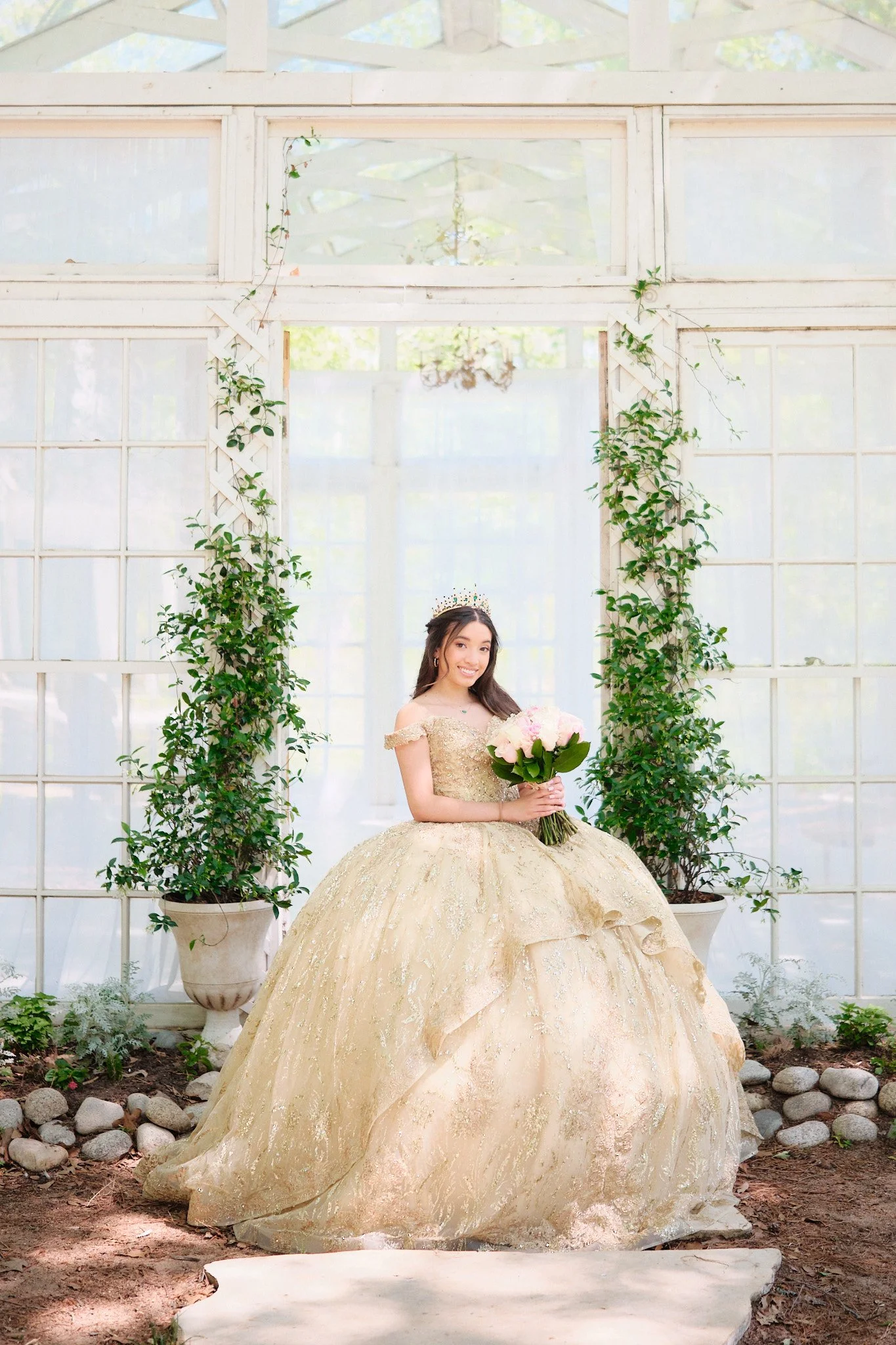 A woman wearing a gold ball gown with a full skirt and off-the-shoulder sleeves, holding a bouquet of pink and white roses, standing in a greenhouse with white wooden walls and green potted plants around her.
