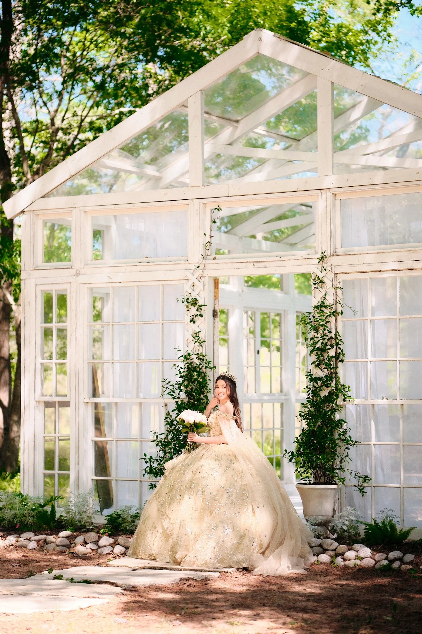 A bride in a gold wedding gown holding a bouquet, standing in front of a white greenhouse with greenery and flowers.