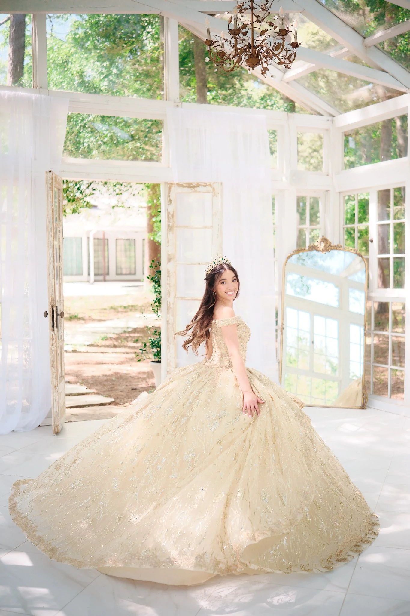 A young woman in a gold ball gown with off-the-shoulder sleeves and a full, elaborate skirt is standing indoors near a large mirror, smiling. The room has large windows, white curtains, and a chandelier hanging from the ceiling, with trees visible ou