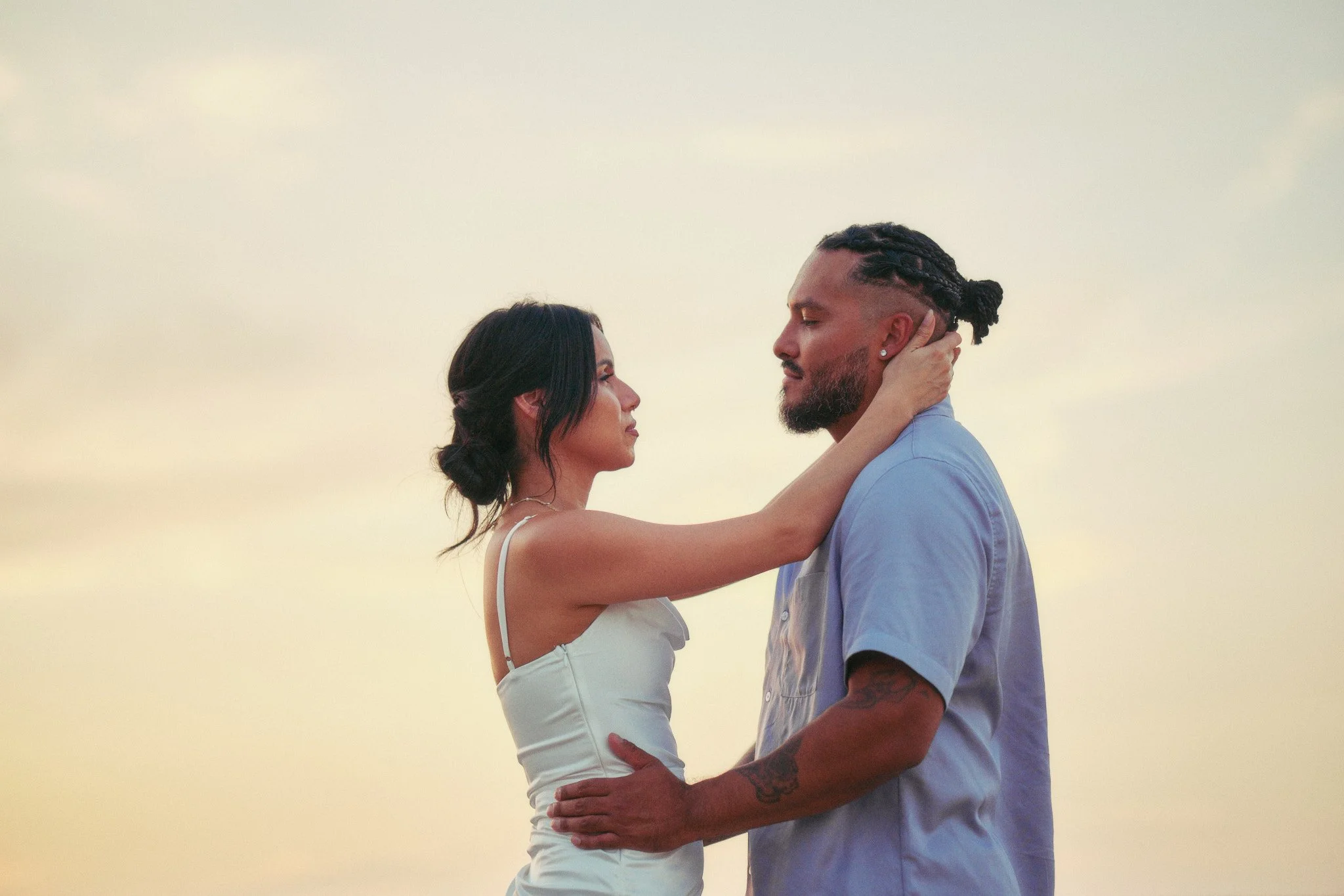 A couple stands close together outdoors on a cloudy day, gazing into each other's eyes, with the woman gently holding the man's face and the man holding her waist.
