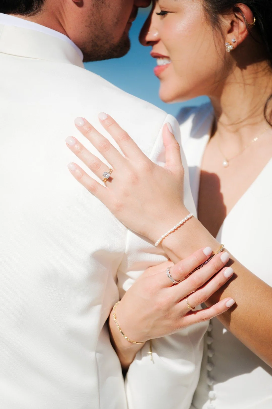 Close-up of a bride and groom embracing, showing the bride's hand with an engagement ring and wedding band, on the groom's shoulder. The bride is smiling, wearing pearl jewelry, and has her arm around the groom.