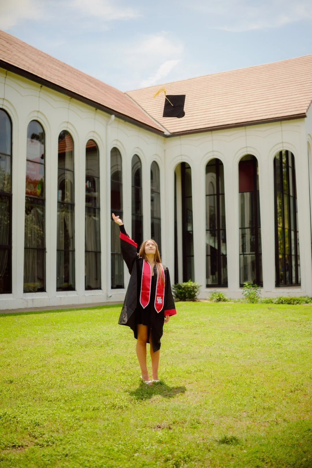 A woman in graduation gown and stole throws her graduation cap into the air on a grassy lawn in front of a modern building with tall windows.