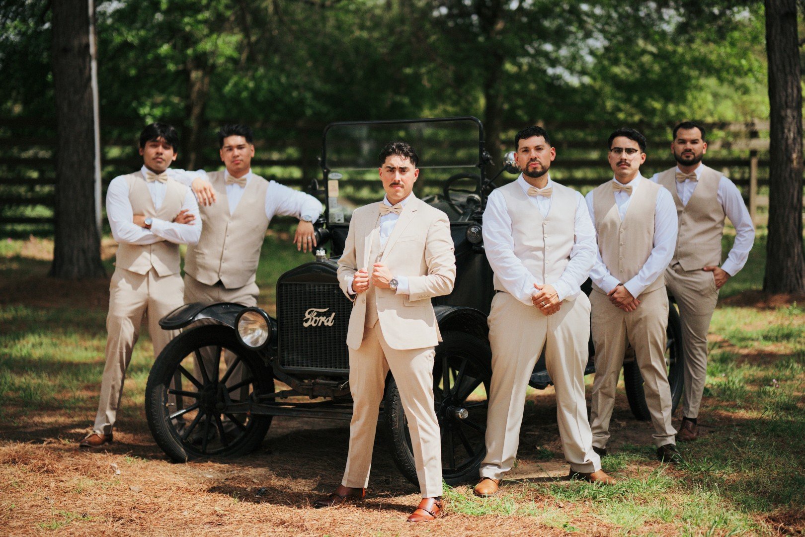 Group of seven men in beige and white suits standing outdoors in front of a vintage black Ford car, with a wooded background.