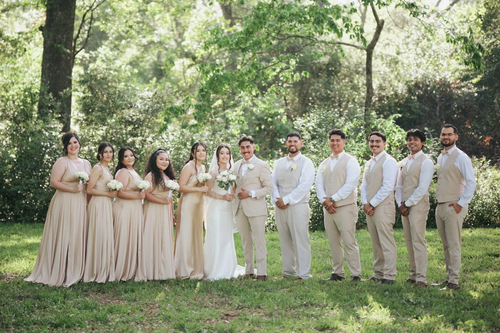 Group portrait of a bride, groom, bridesmaids in beige dresses, and groomsmen in beige vests and pants, standing outdoors on grass with trees in the background, during a wedding celebration.
