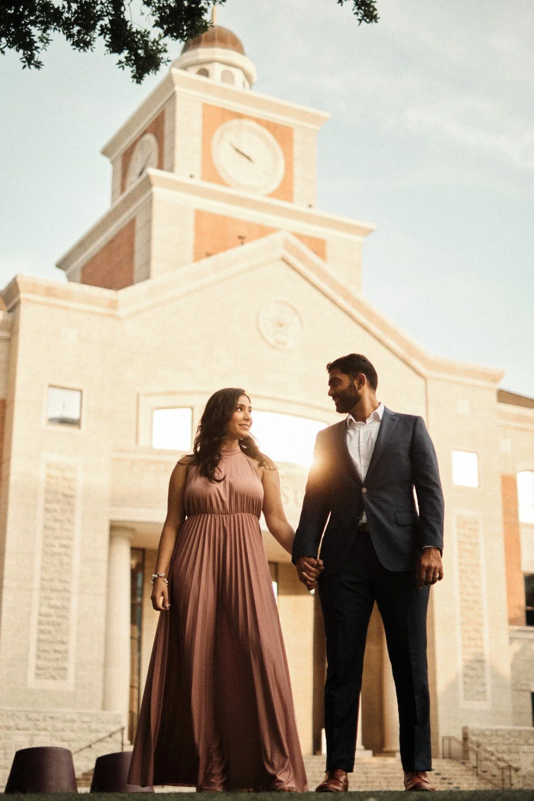 A couple holding hands and walking in front of a large building with a clock tower, during sunset, with trees in the background.