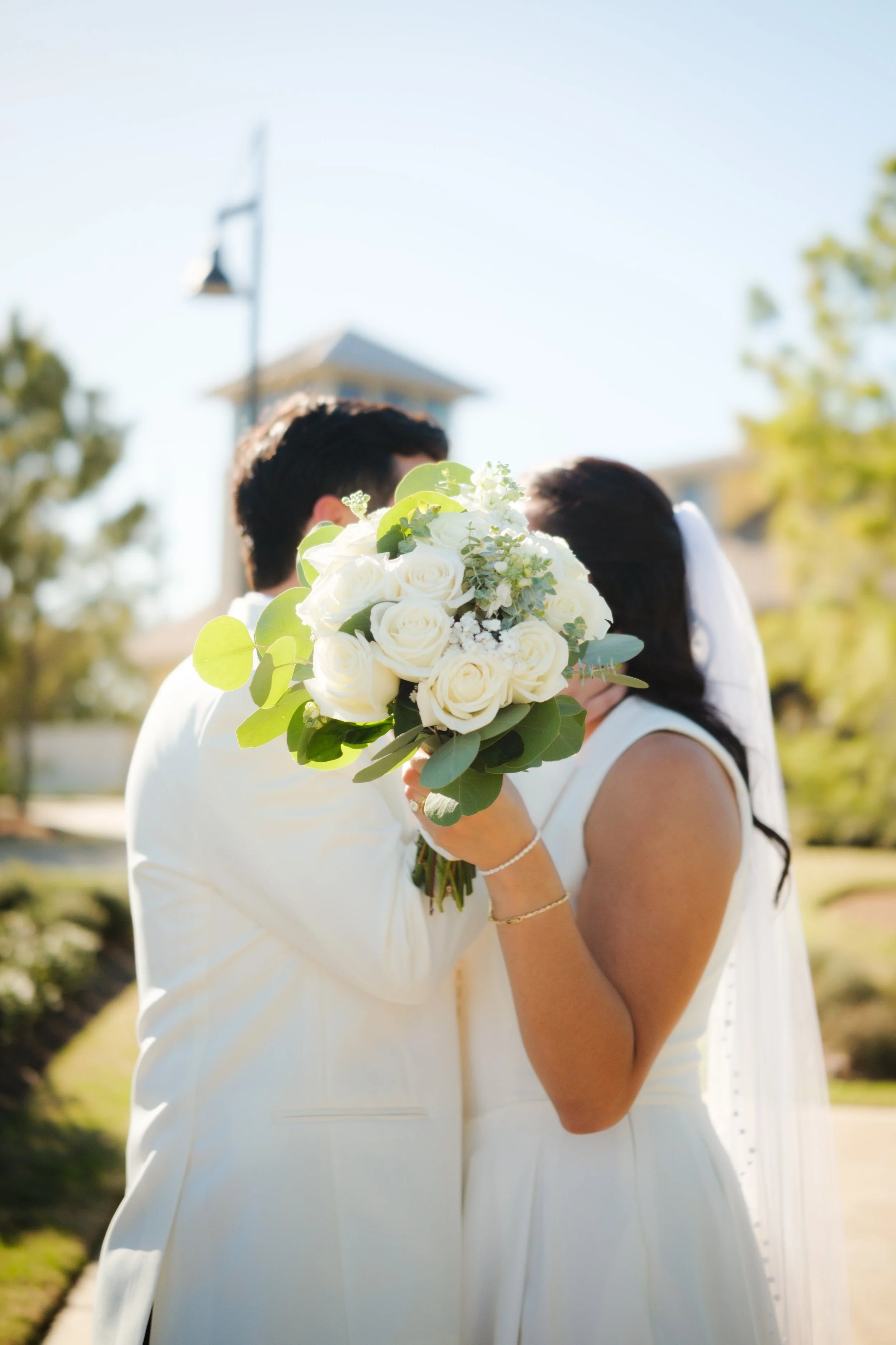 A bride and groom are holding a bouquet of white roses and greenery, standing close together outdoors with a church and trees in the background on a sunny day.