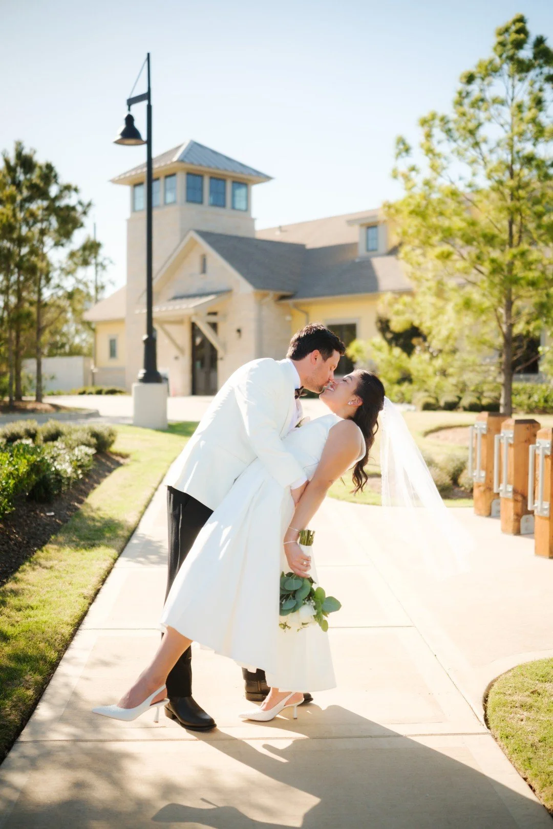 A bride and groom sharing a kiss outside a church on their wedding day, with the groom dipping the bride slightly. The bride is holding a bouquet of eucalyptus leaves, and both are dressed in white wedding attire. The scene is sunny with a clear sky,