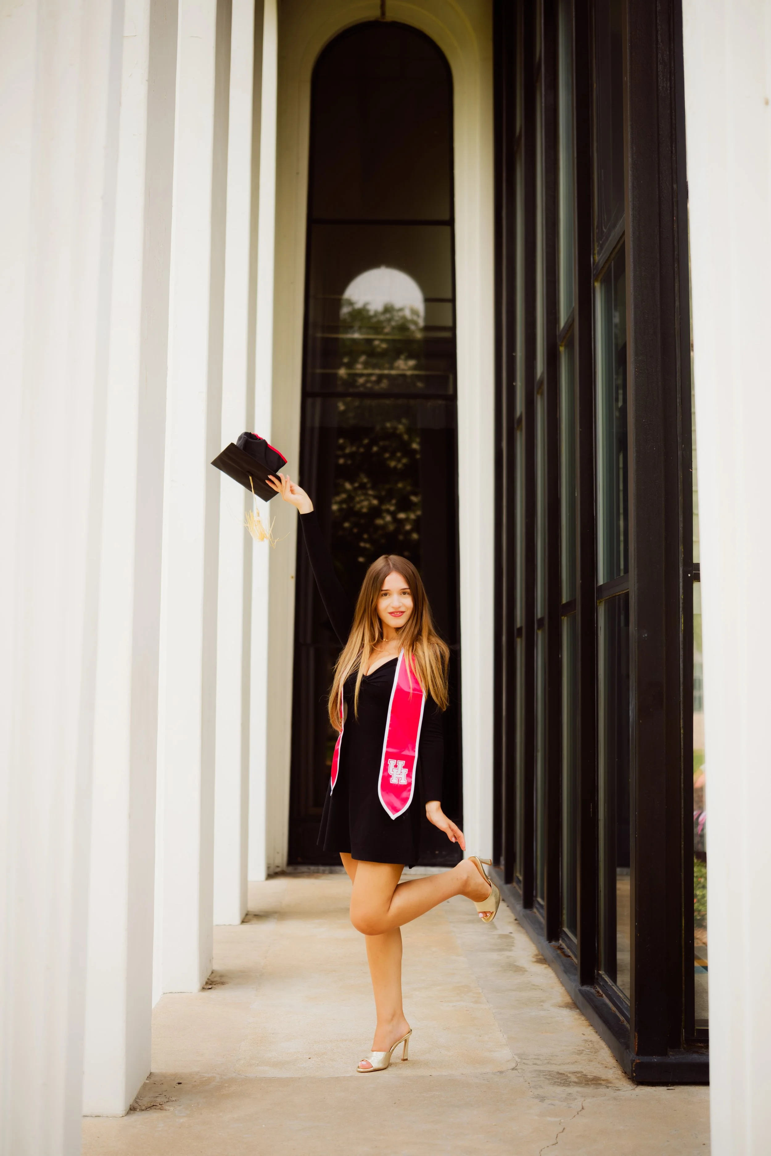 A young woman in a black dress and gold high heels is happily jumping with one leg bent and the other extended, holding a black graduation cap in one hand. She is standing in a hallway with white vertical paneling on the left and large glass windows 