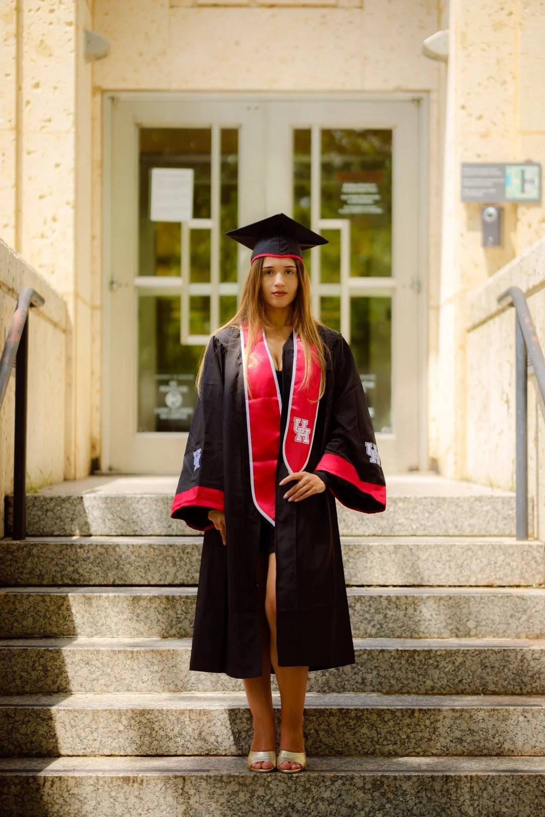 A young woman in a black graduation gown and cap with a red and white stole, standing on university steps in front of a glass door.