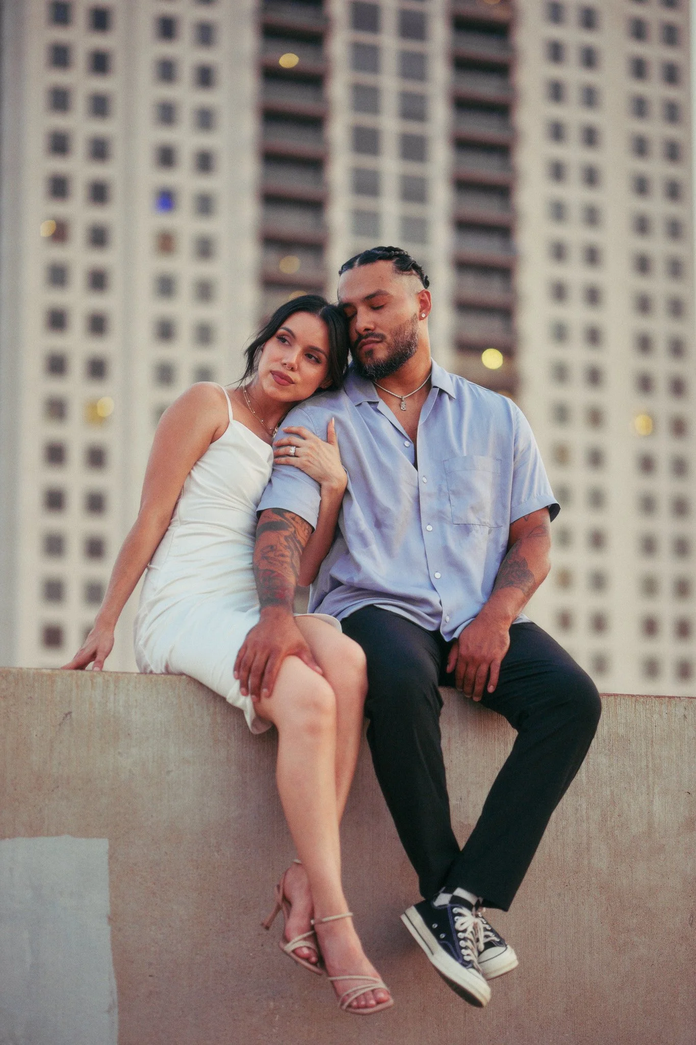 A man and a woman sitting close together outdoors in front of a tall building. The woman has her head resting on the man's shoulder, and they both have relaxed, peaceful expressions. The woman is wearing a white dress and heels, while the man is in a