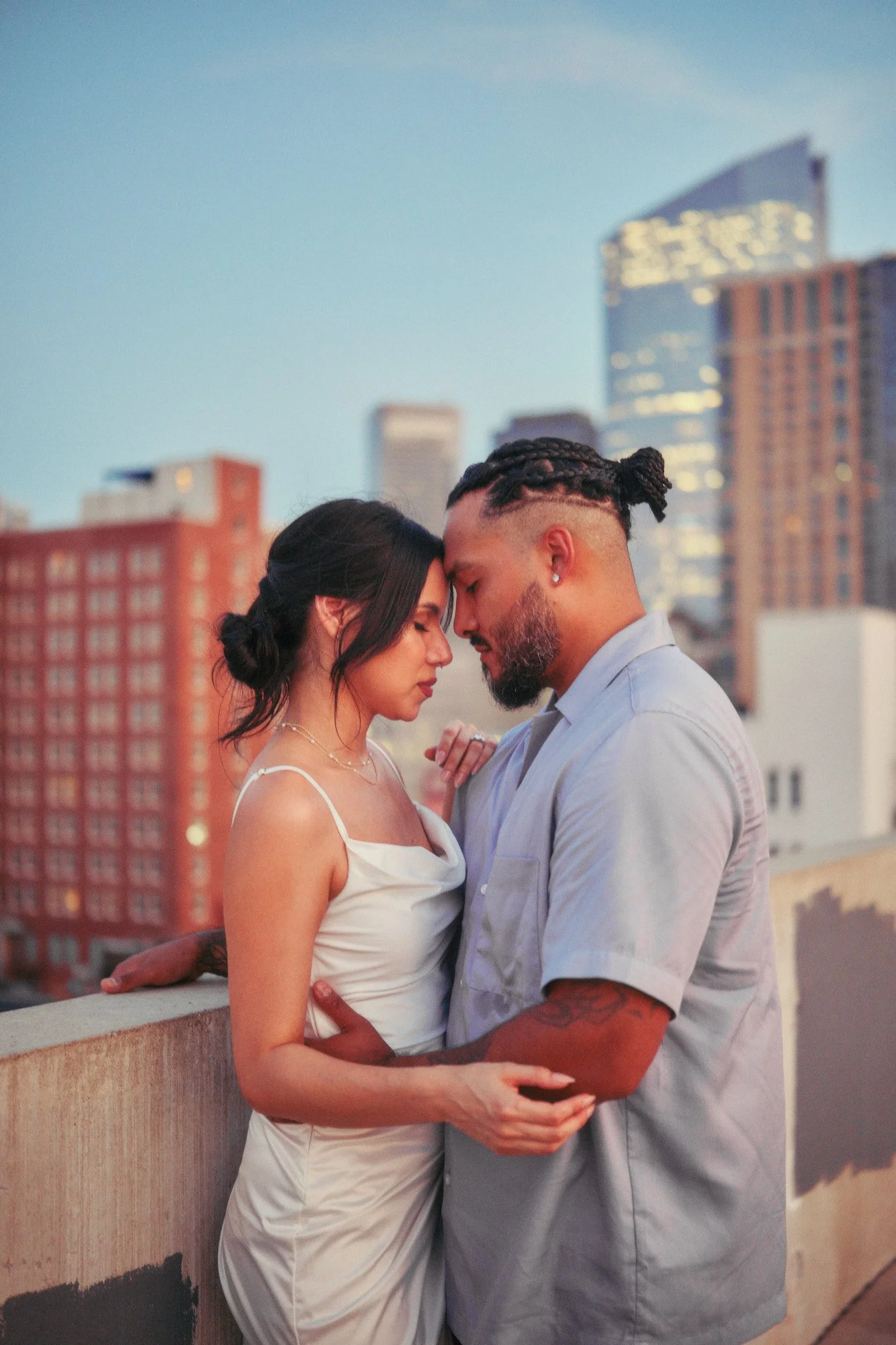 A couple standing close together on a rooftop with a city skyline in the background during dusk, with their foreheads touching and eyes closed.