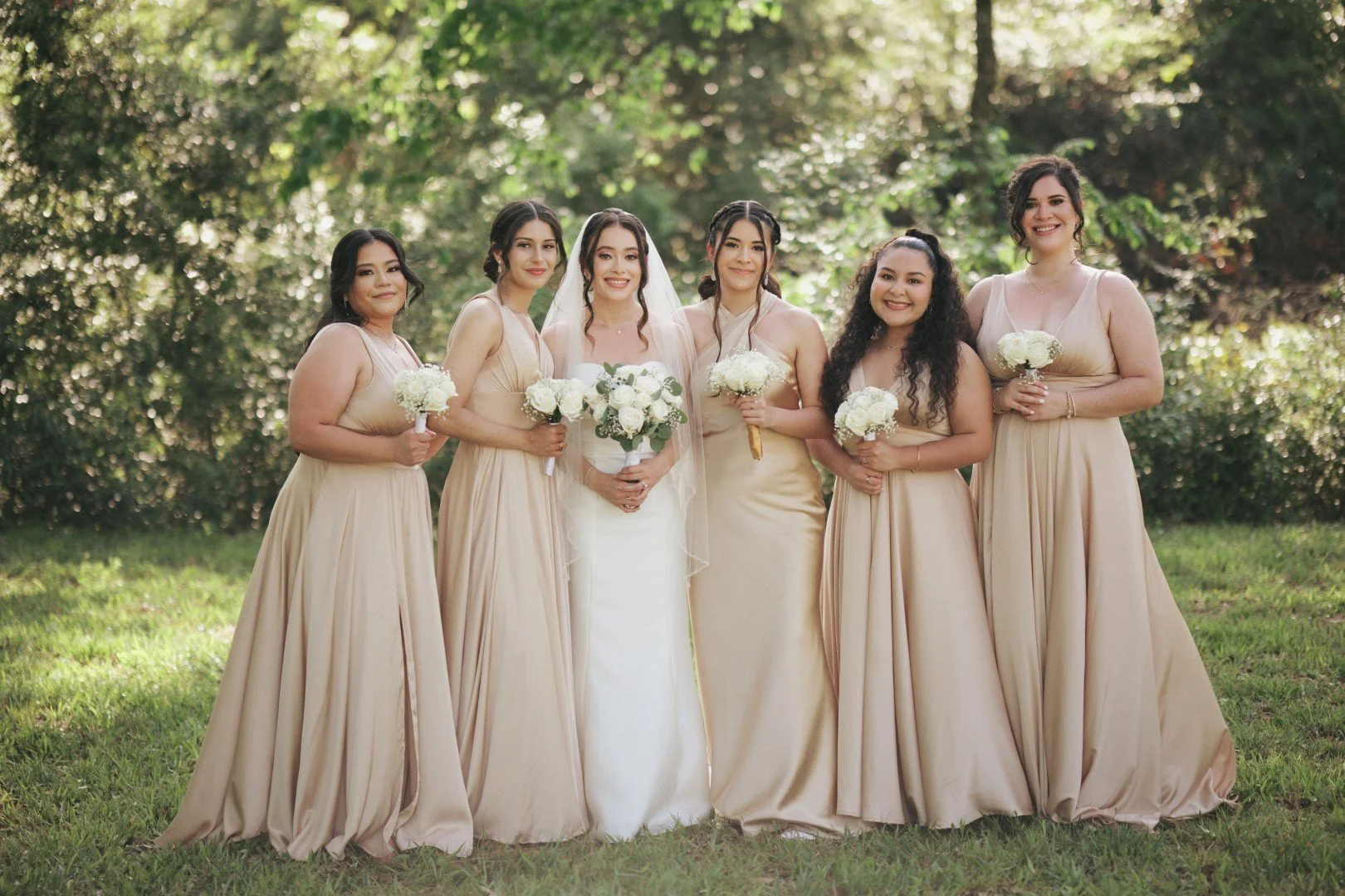 A group of six women standing outdoors in front of trees, dressed in matching beige bridesmaid dresses, holding white flower bouquets, with a bride in a white gown and veil in the center, all smiling.