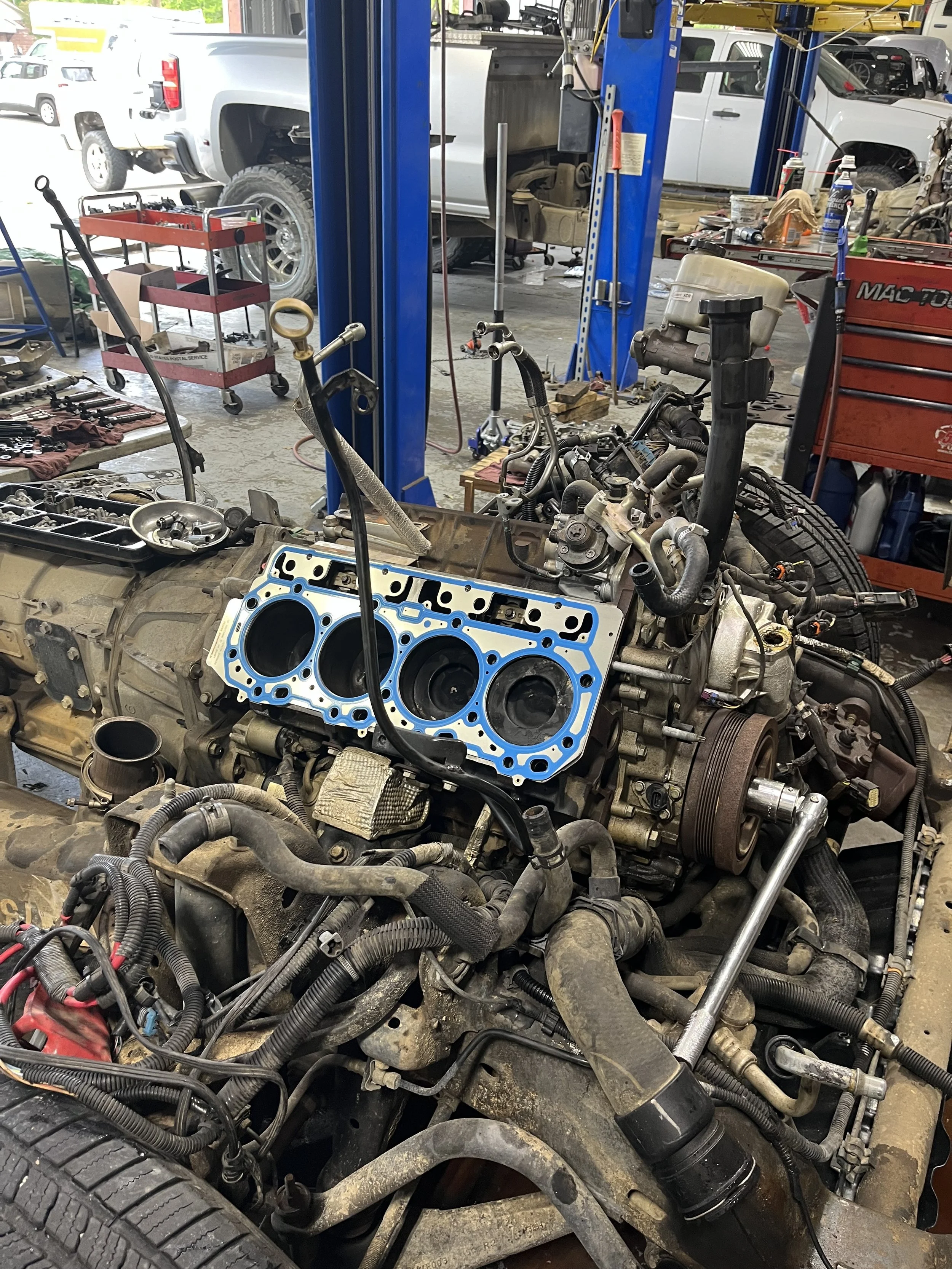 View of an engine in a repair shop, with various tools and car parts scattered around. A vehicle is elevated on a lift in the background, while the engine is partially disassembled with hoses, wires, and mechanical components visible.