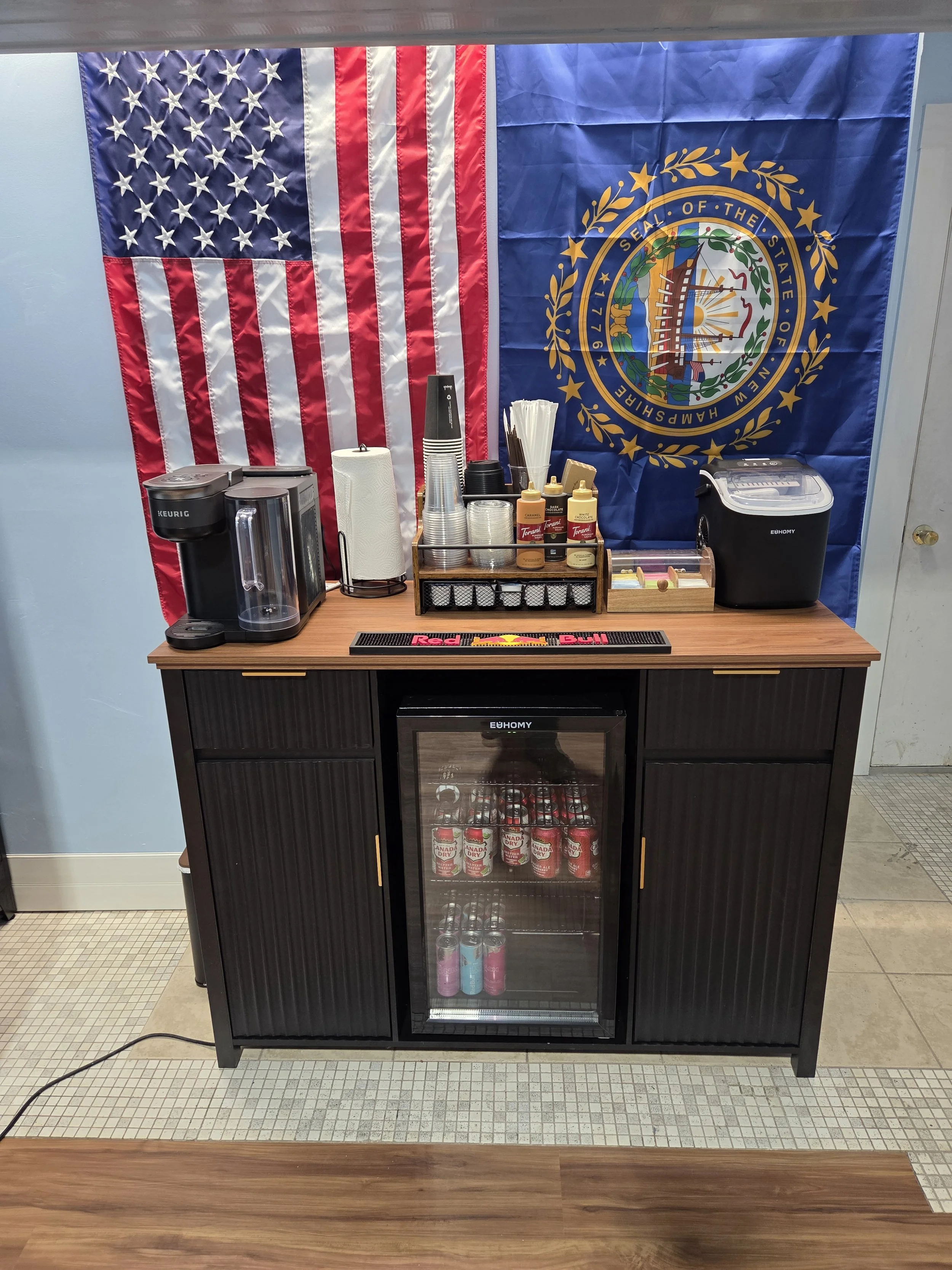 A beverage station setup with an American flag and a Maine state flag hanging on the wall. The station includes a Keurig coffee maker, disposable cups, condiments, straws, and a small refrigerator stocked with cans of soda.