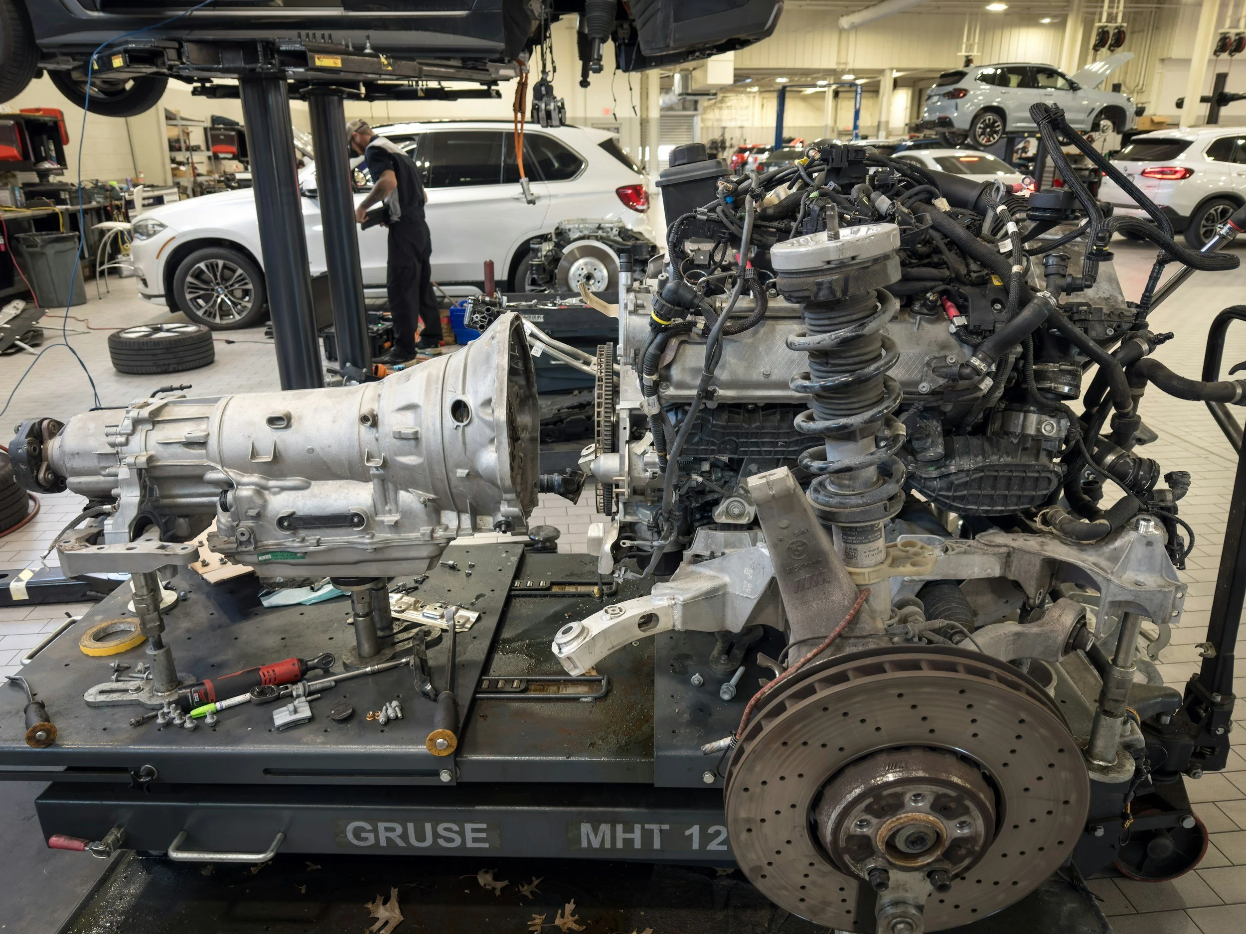 An automotive repair shop with a large engine and transmission removed from a vehicle. Various tools and parts are scattered on a workbench. In the background, a mechanic is working on a white car that is elevated on a lift, with additional cars parked or being serviced in the spacious garage.
