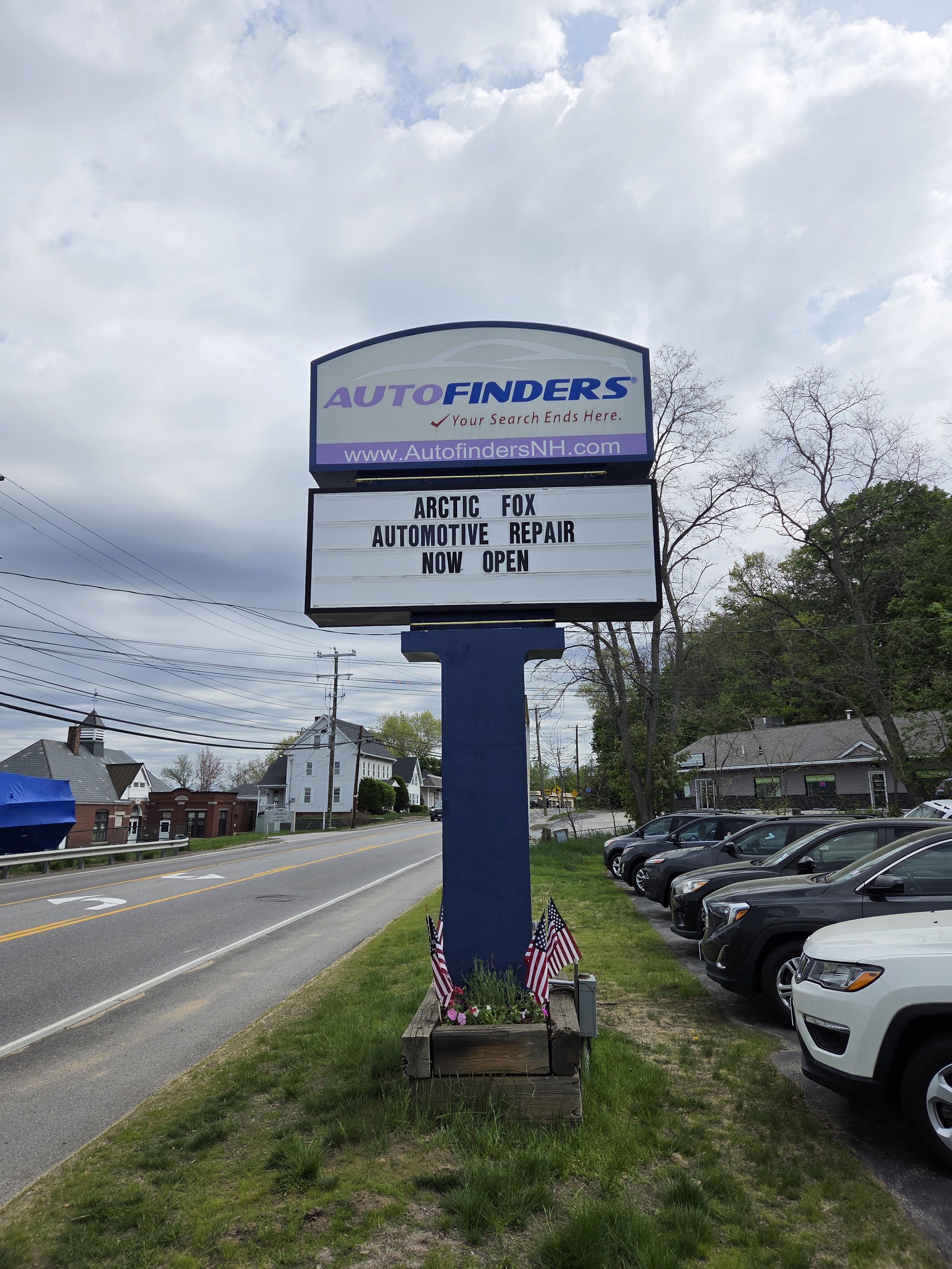 A roadside sign advertising Arctic Fox Automotive Repair, now open, with American flags and flowers at the base, along a street with parked cars and houses in the background.