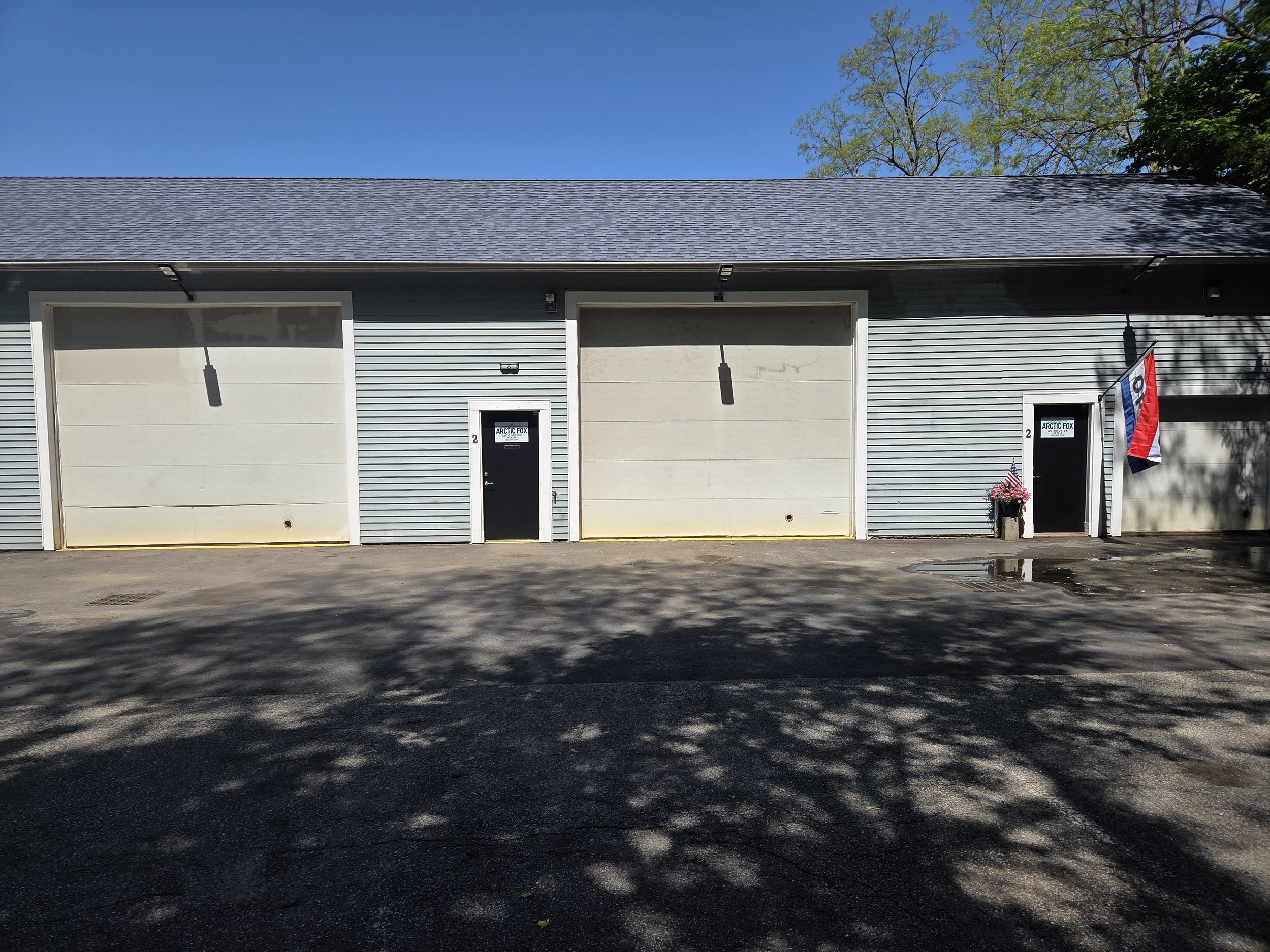 A photo of a gray building with two garage doors, two small doors, and a small flag with red, white, and blue colors. There are trees and a clear blue sky in the background.