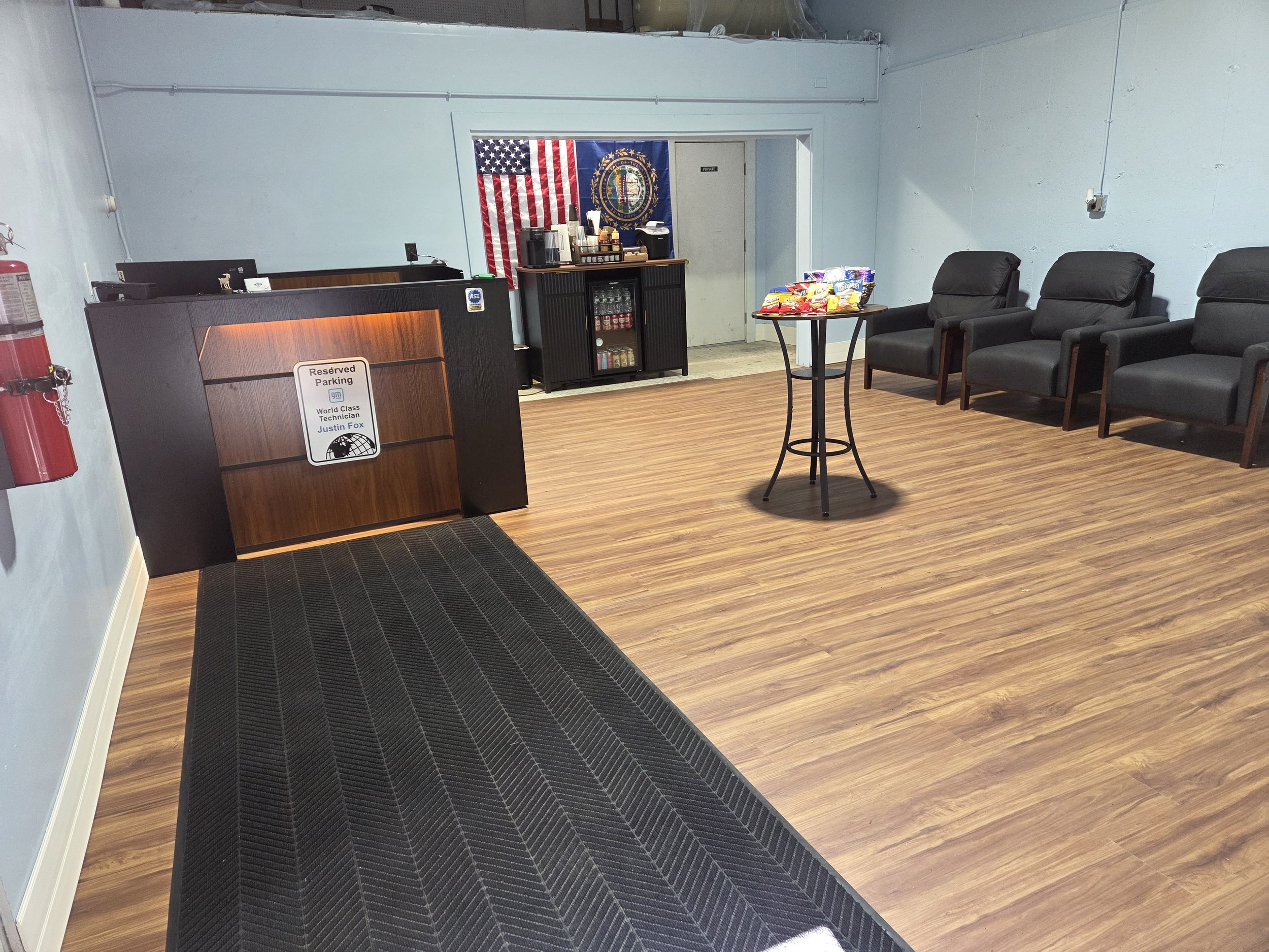 An indoor waiting area with four black chairs with armrests, a small round table with snack bags, a black reception desk with a reserved parking sign, and a snack station behind the desk. The walls are light blue, and there is a fire extinguisher on the left. An American flag and a state flag hang on the wall behind the snack station.