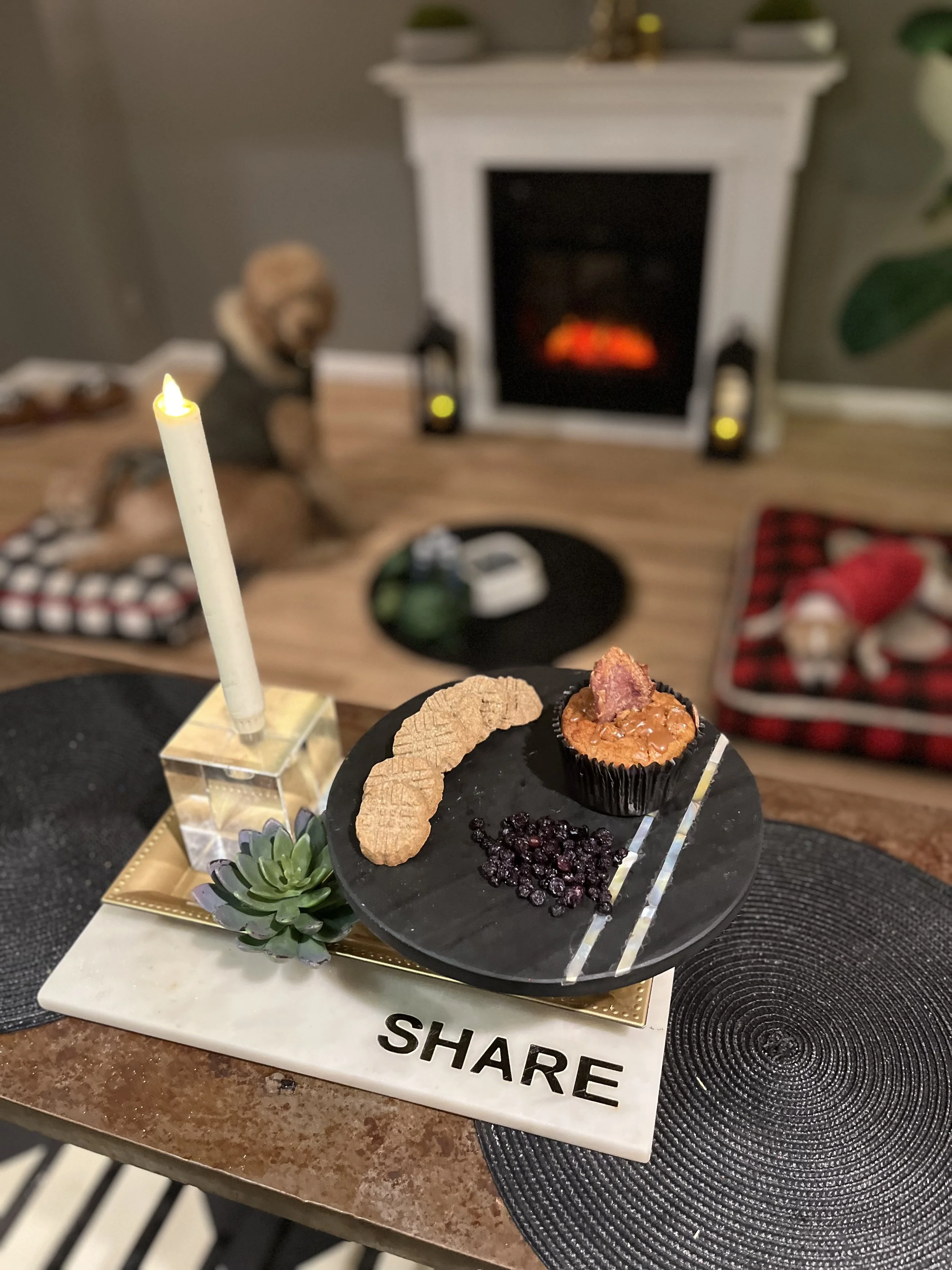 A black plate with a cupcake, cookies, blueberries, and a piece of bacon on a white board that has the words 'SHARE' in black. A white candle with a yellow flame sits next to the plate. The background shows a living room with a fireplace, lanterns, and a person sitting on the floor.