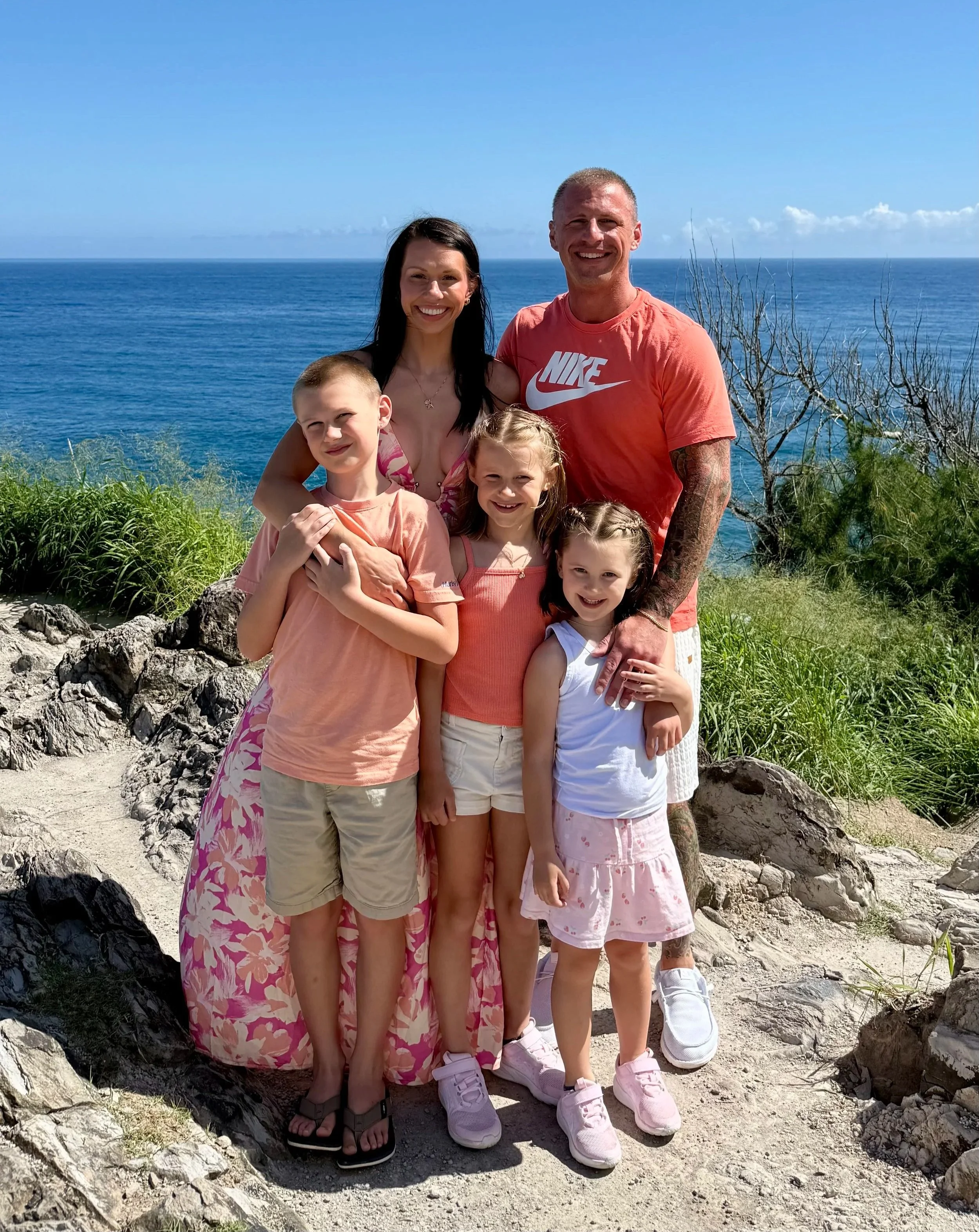 A family of six, including two adults and four children, standing close together on a rocky path with an ocean in the background under a clear blue sky.